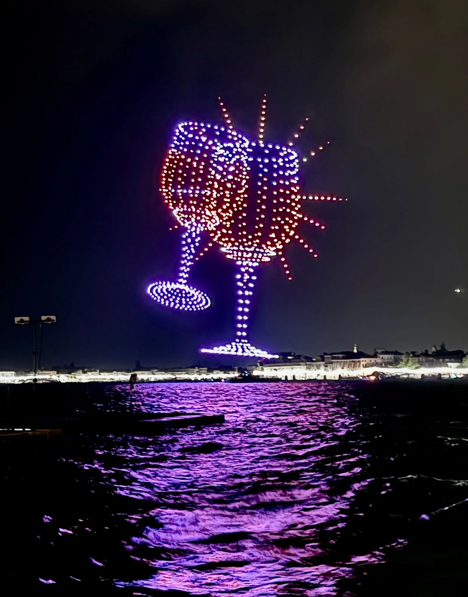 Due bicchieri che brindano disegnati dai droni nel cielo notturno, con un'esplosione di luce intorno – Aperol Redentore Drone Show, Venezia, Italia, 2024 – direzione creativa