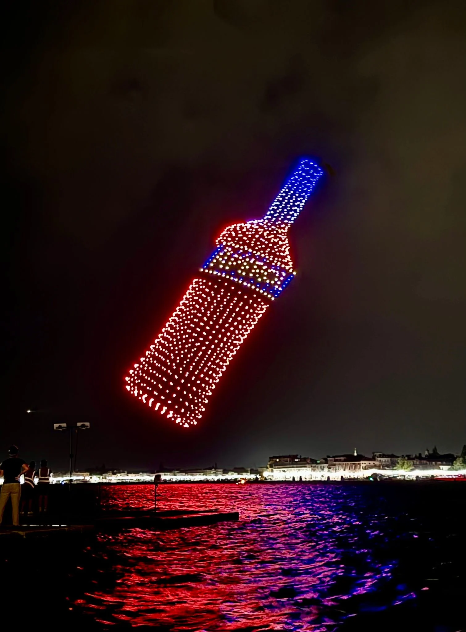 Bottiglia di Aperol inclinata disegnata da droni rossi e blu, vista dalla riva con la laguna illuminata sotto – Aperol Redentore Drone Show, Venezia, Italia, 2024 – direzione creativa