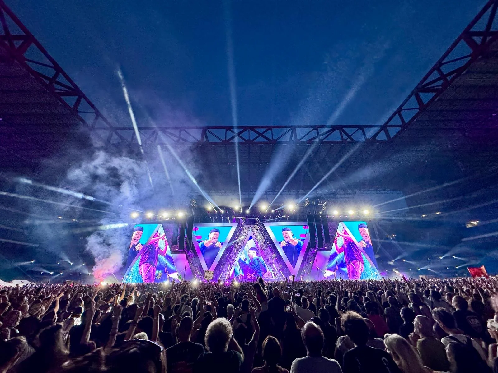 Vista notturna del palco con maxischermi triangolari, fasci di luce e fumo che sale sullo stadio gremito – Vasco Rossi Live 2024, Italia – scenografia per concerti