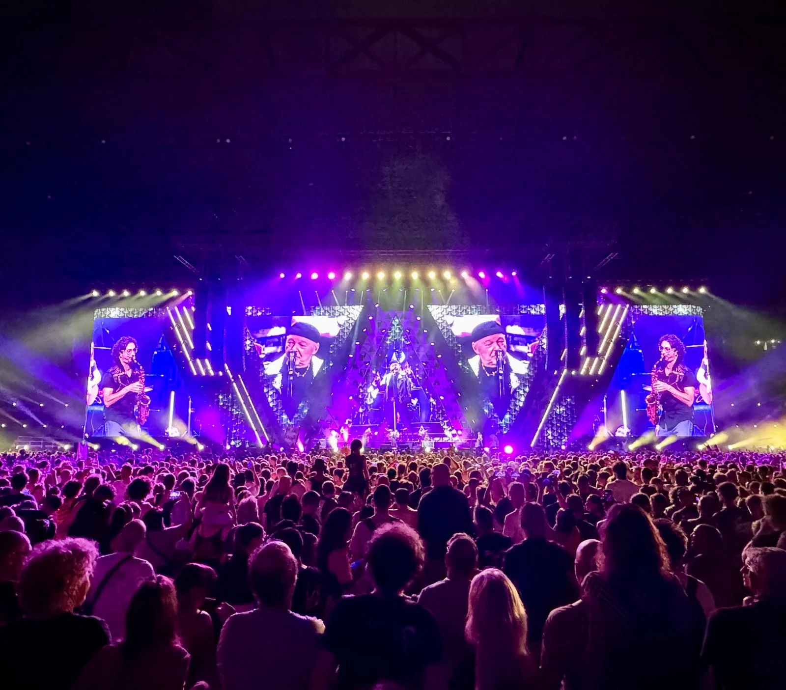 Palco immerso in luce viola, schermi con un sassofonista e il cantante mentre la folla guarda – Vasco Rossi Live 2024, Italia – scenografia per concerti