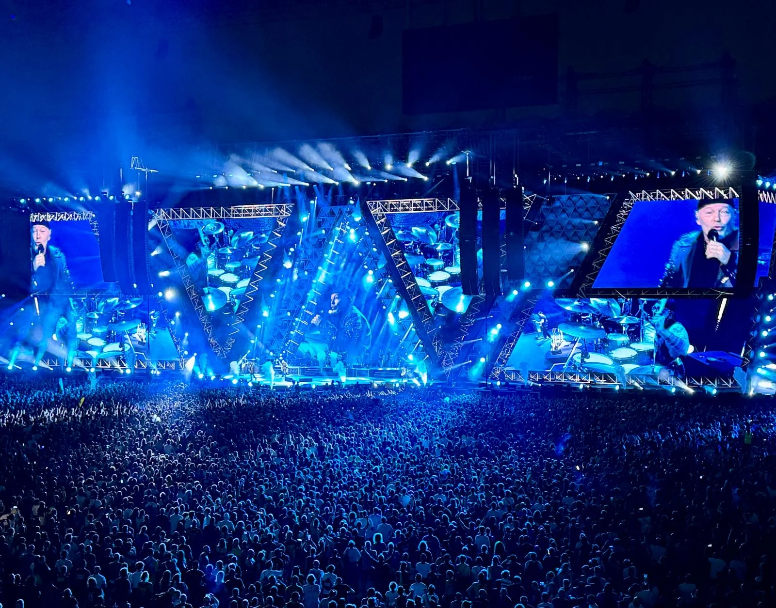 Palco e folla in luce blu intensa, telai angolari di americane e schermi con primi piani del cantante – Vasco Rossi Live 2024, Italia – scenografia per concerti