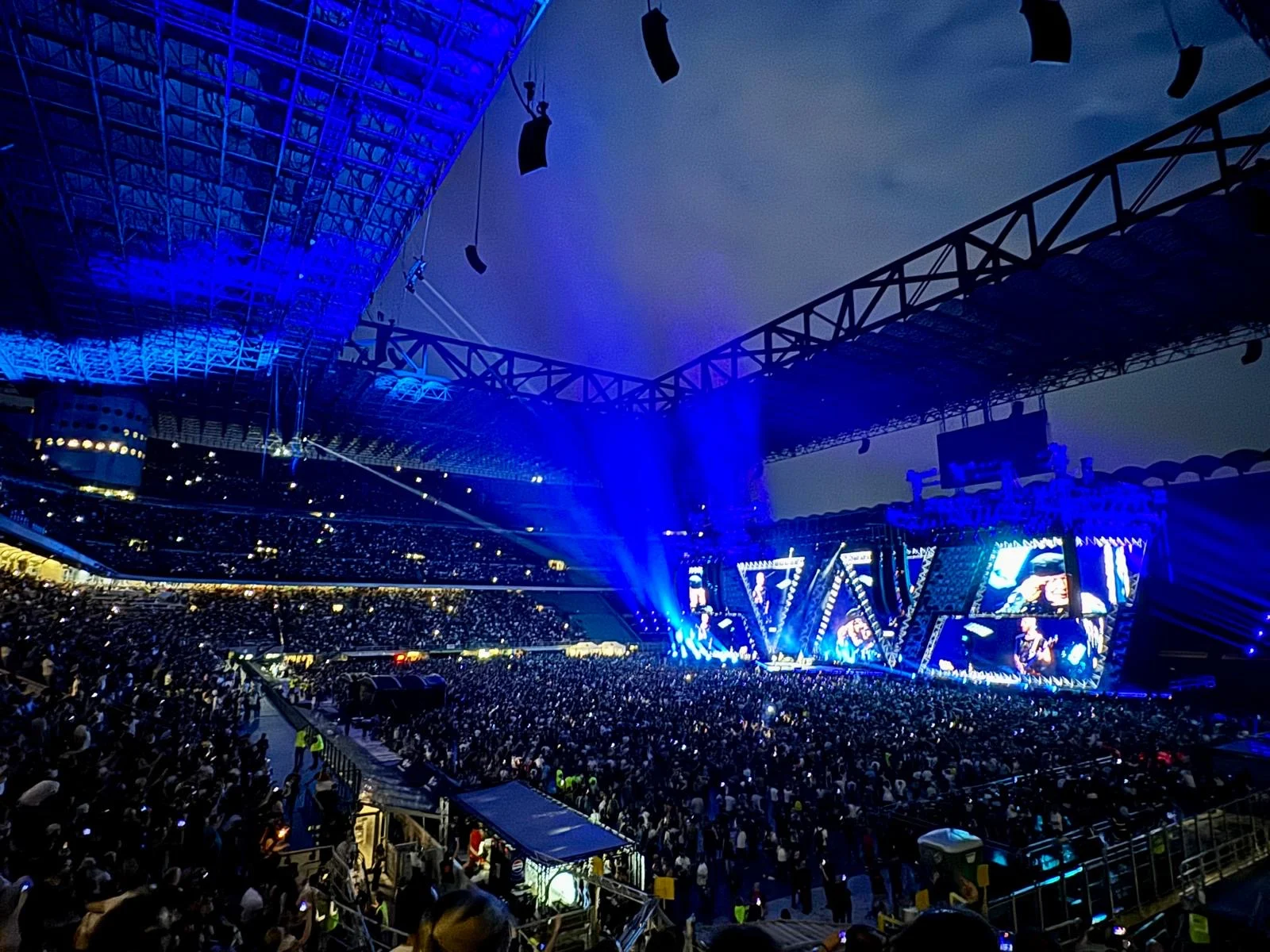 Vista dagli spalti superiori: stadio illuminato di blu, folla sul prato e gli schermi del palco in lontananza – Vasco Rossi Live 2024, Italia – scenografia per concerti