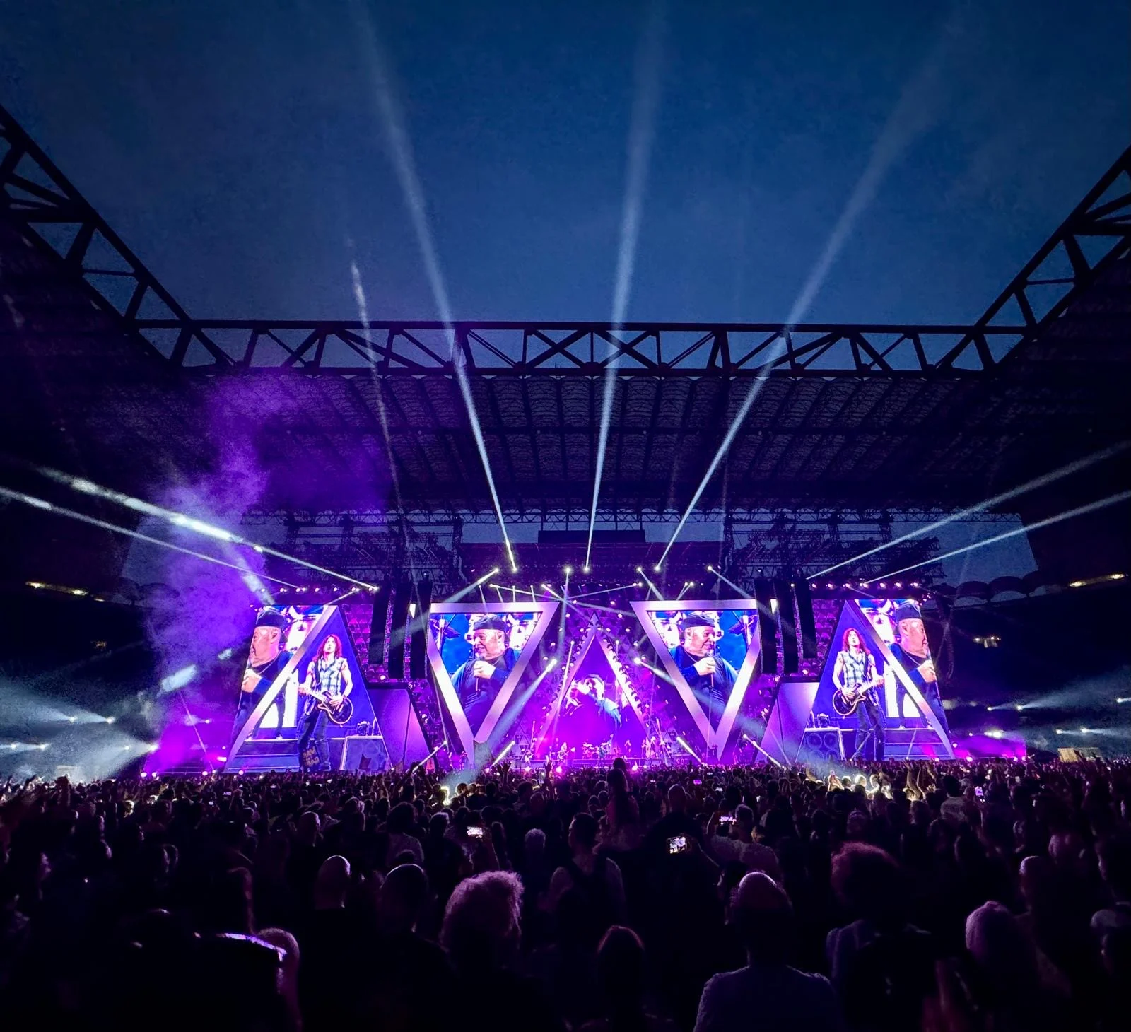 Palco illuminato di viola con fasci di fari che spazzano il cielo notturno e fumo che si muove sulla folla – Vasco Rossi Live 2024, Italia – scenografia per concerti