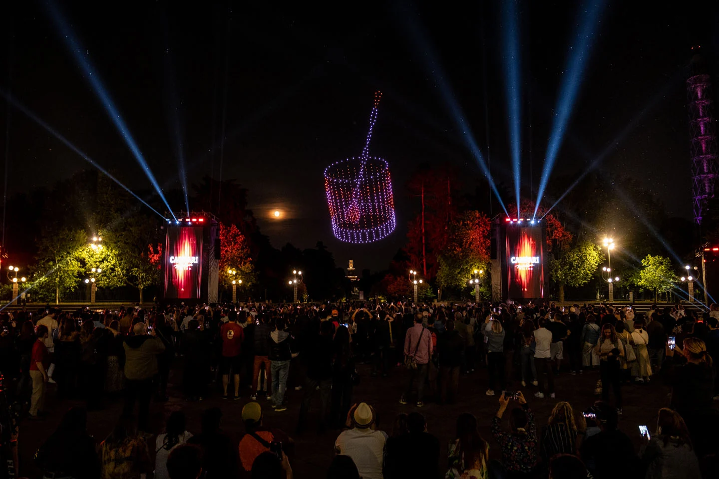 Droni che formano un cesto luminoso sopra la folla di notte, fari e torri rosse Campari ai lati – Milano House of Campari, Italia, 2023 – progettazione di eventi