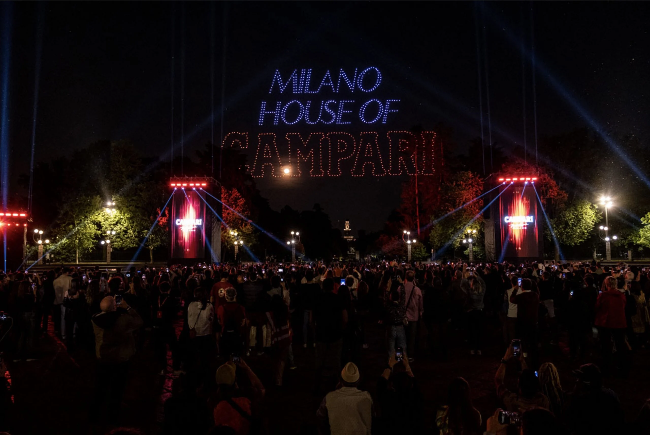 Droni che compongono la scritta Milano House of Campari nel cielo notturno sopra la folla e le torri Campari illuminate di rosso – Milano House of Campari, Italia, 2023 – progettazione di eventi