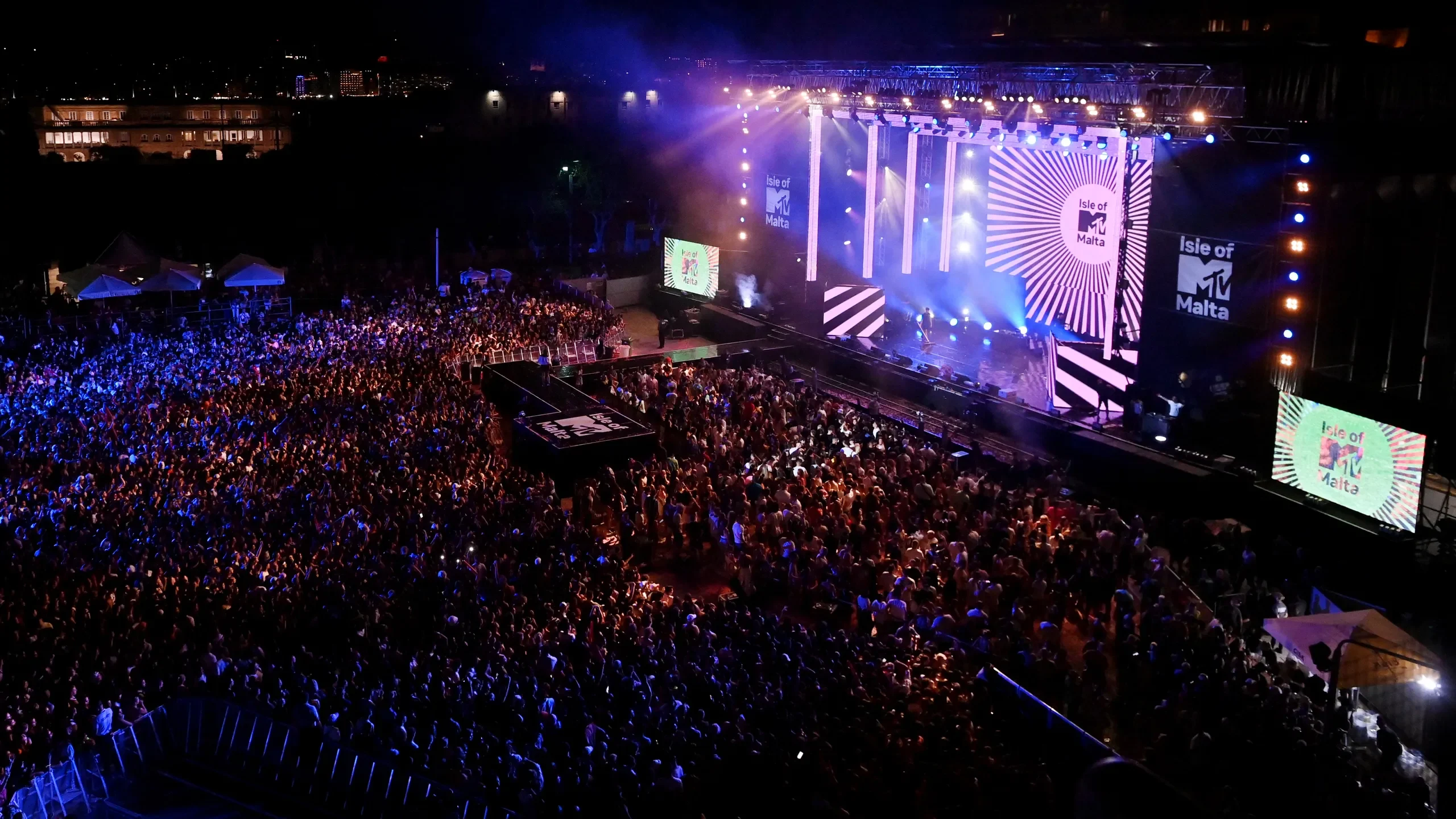 Vista aerea della piazza di Floriana gremita davanti al palco di Isle of MTV, schermi laterali e raggi di luce bianca – Isle of MTV 2024, Floriana, Malta – scenografia per concerti