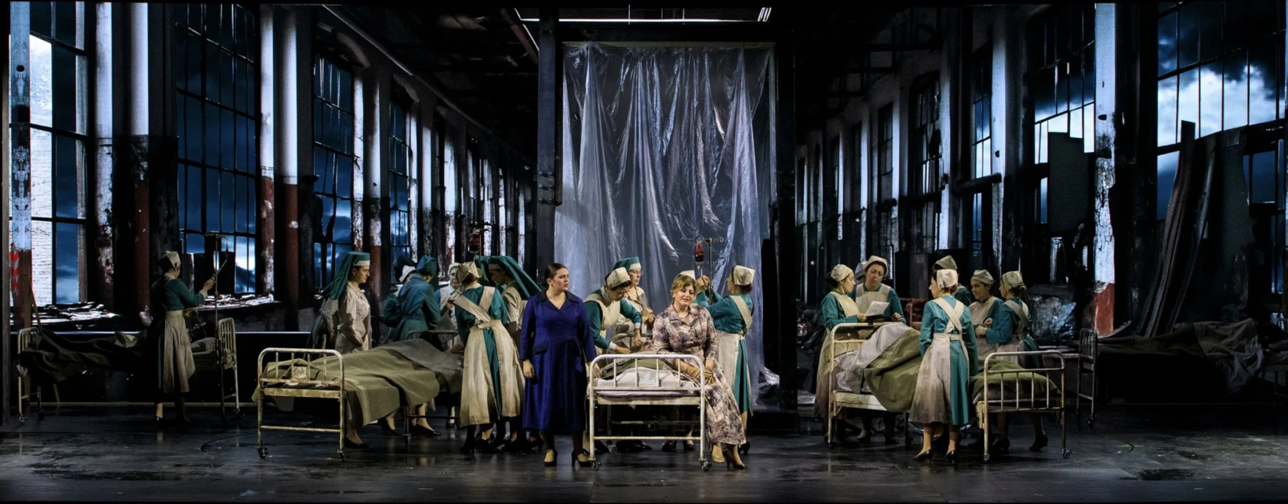Infermiere in divise chiare intorno a letti d'ospedale in un capannone industriale abbandonato con alte finestre – Il Trovatore al Teatro Comunale Nouveau, Bologna, Italia, 2024 – scenografia d'opera