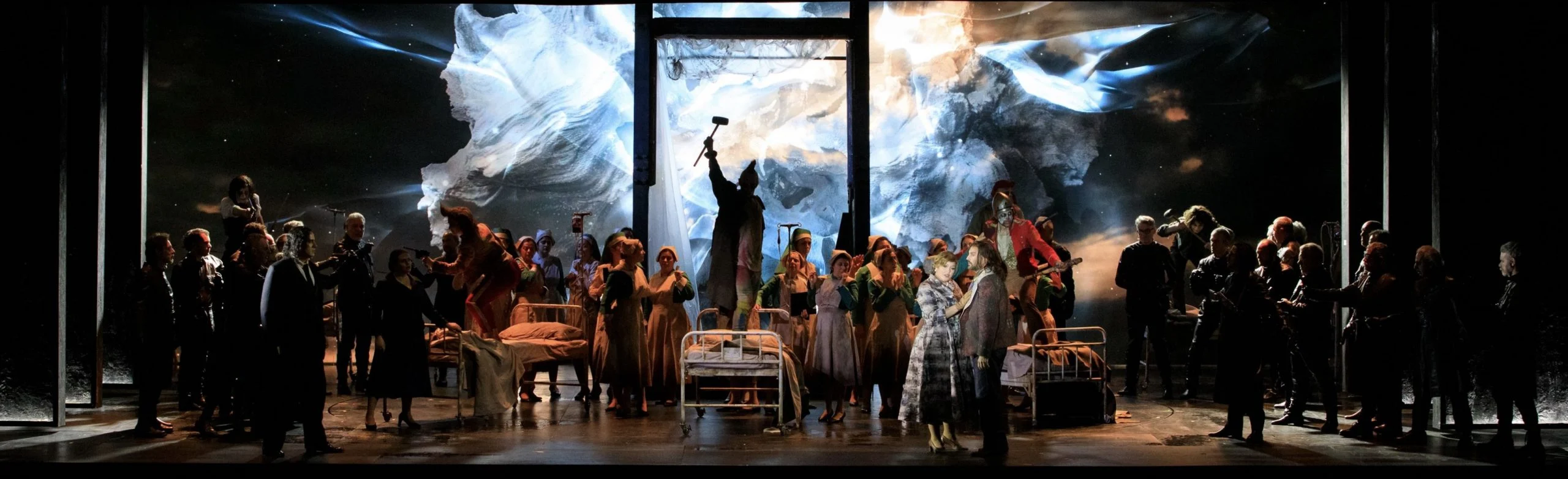 Coro intorno a letti d'ospedale su un palco buio, con una tempesta di ghiaccio bianca proiettata su tutto il fondale – Il Trovatore al Teatro Comunale Nouveau, Bologna, Italia, 2024 – scenografia d'opera