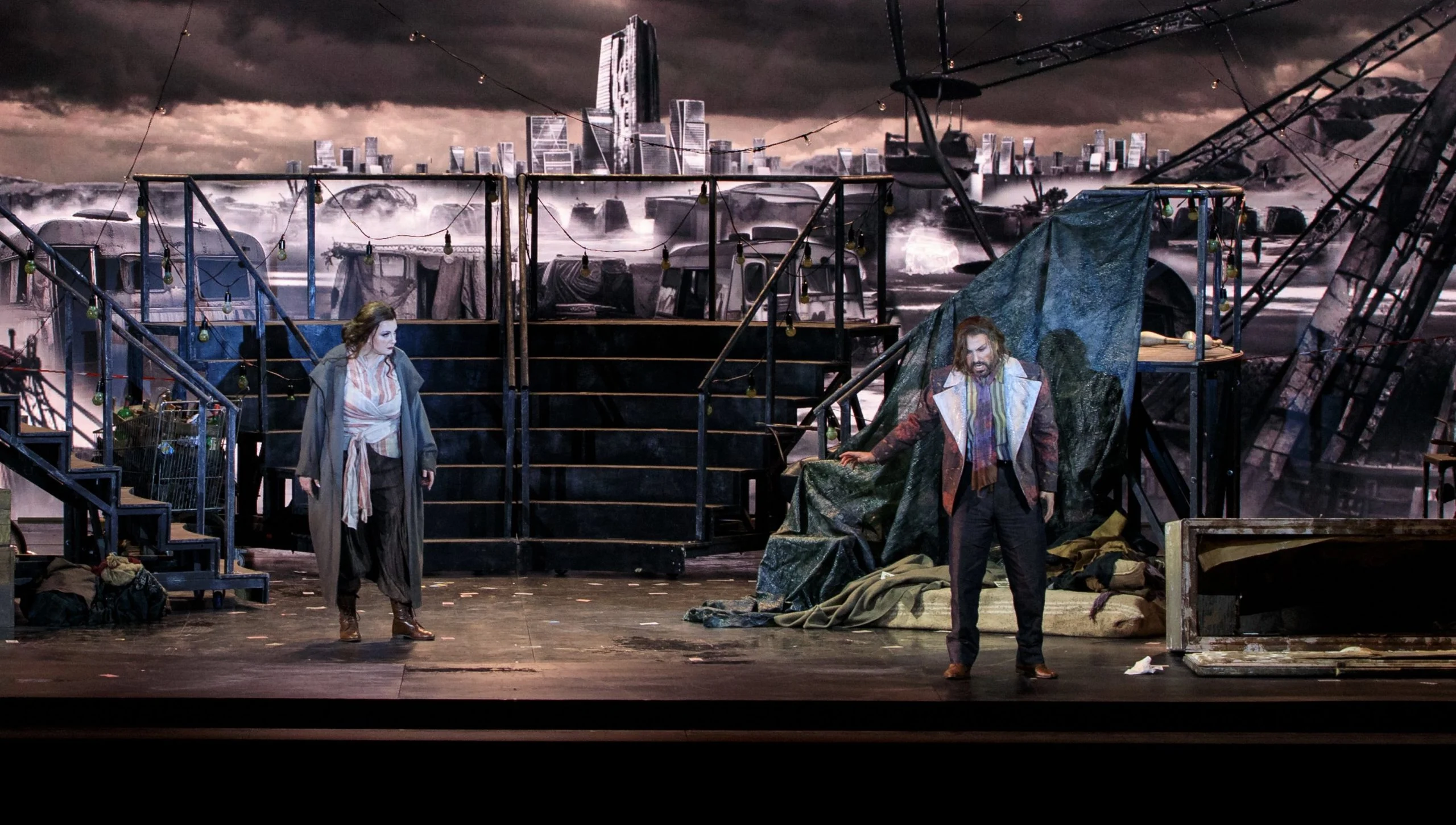 Due cantanti uno di fronte all'altro su un palco buio con scale metalliche, teloni e lo skyline di una città in rovina dietro – Il Trovatore al Teatro Comunale Nouveau, Bologna, Italia, 2024 – scenografia d'opera