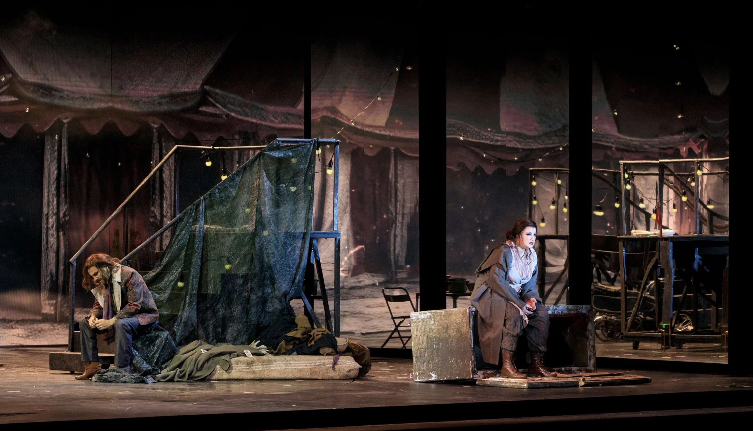 Due cantanti seduti a distanza di notte in una scenografia circense di tende, lucine e impalcature metalliche – Il Trovatore al Teatro Comunale Nouveau, Bologna, Italia, 2024 – scenografia d'opera