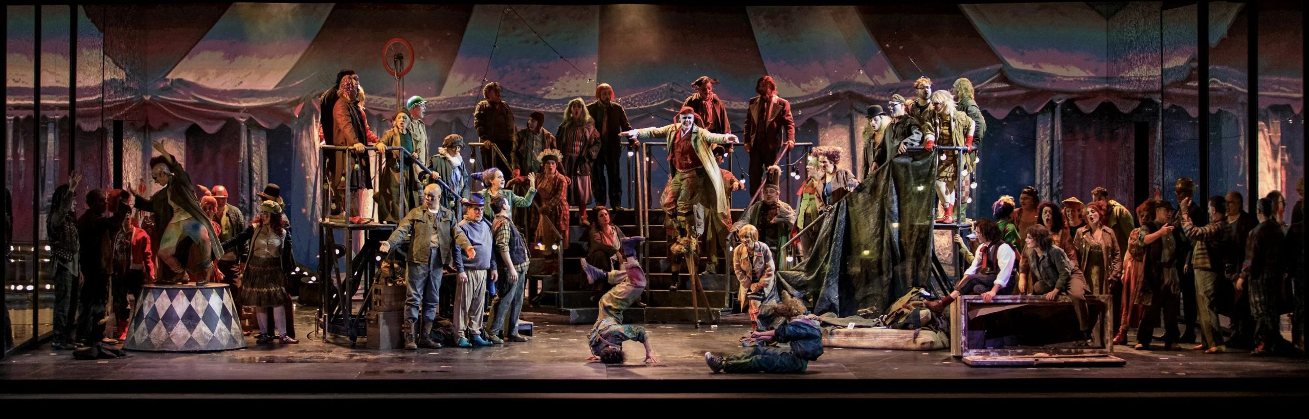 Allestimento de Il Trovatore: un'affollata scena circense con artisti su scale metalliche sotto uno sbiadito tendone da circo – Il Trovatore al Teatro Comunale Nouveau, Bologna, Italia, 2024 – scenografia d'opera