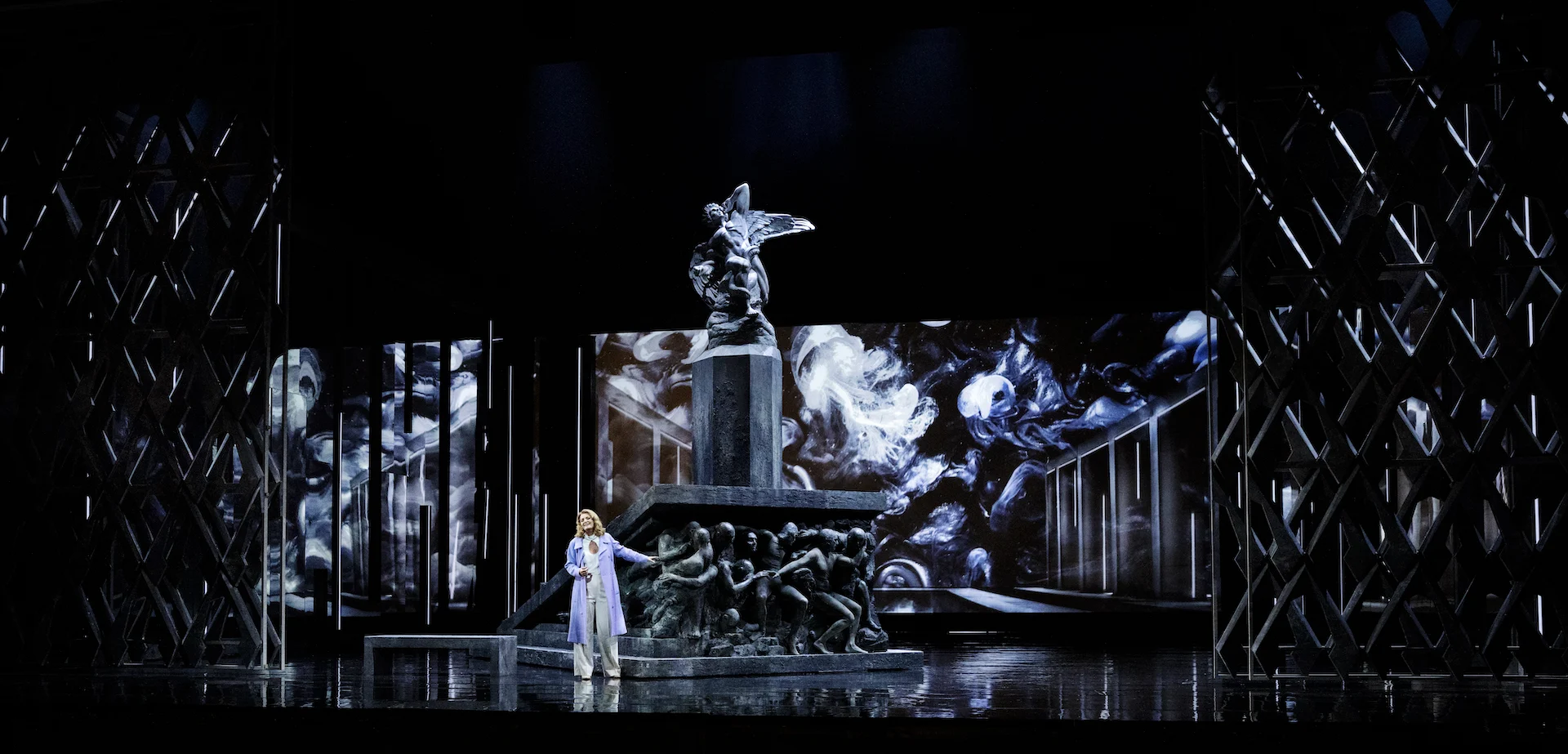 Cantante in bianco davanti a un monumento sormontato da una statua alata, proiezioni monocrome e torri a traliccio – Don Carlo al Teatro Reale Danese, Copenaghen, Danimarca, 2024 – scenografia d'opera