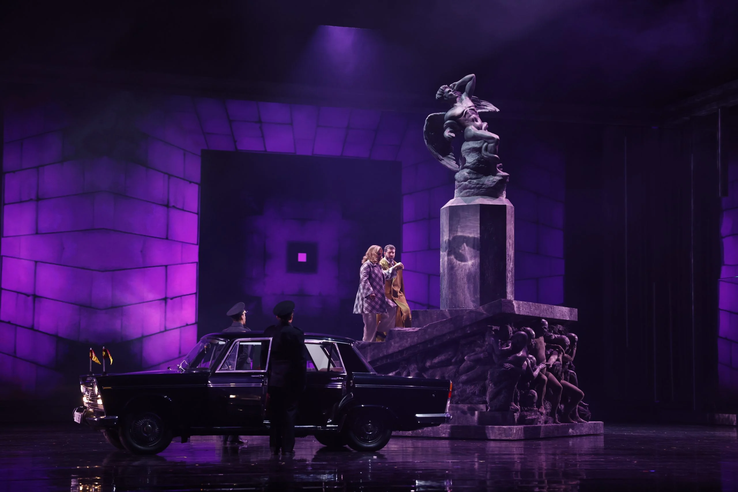 Due cantanti sui gradini di un monumento scolpito immerso in luce viola, un'auto d'epoca nera e guardie sotto – Don Carlo al Teatro Reale Danese, Copenaghen, Danimarca, 2024 – scenografia d'opera