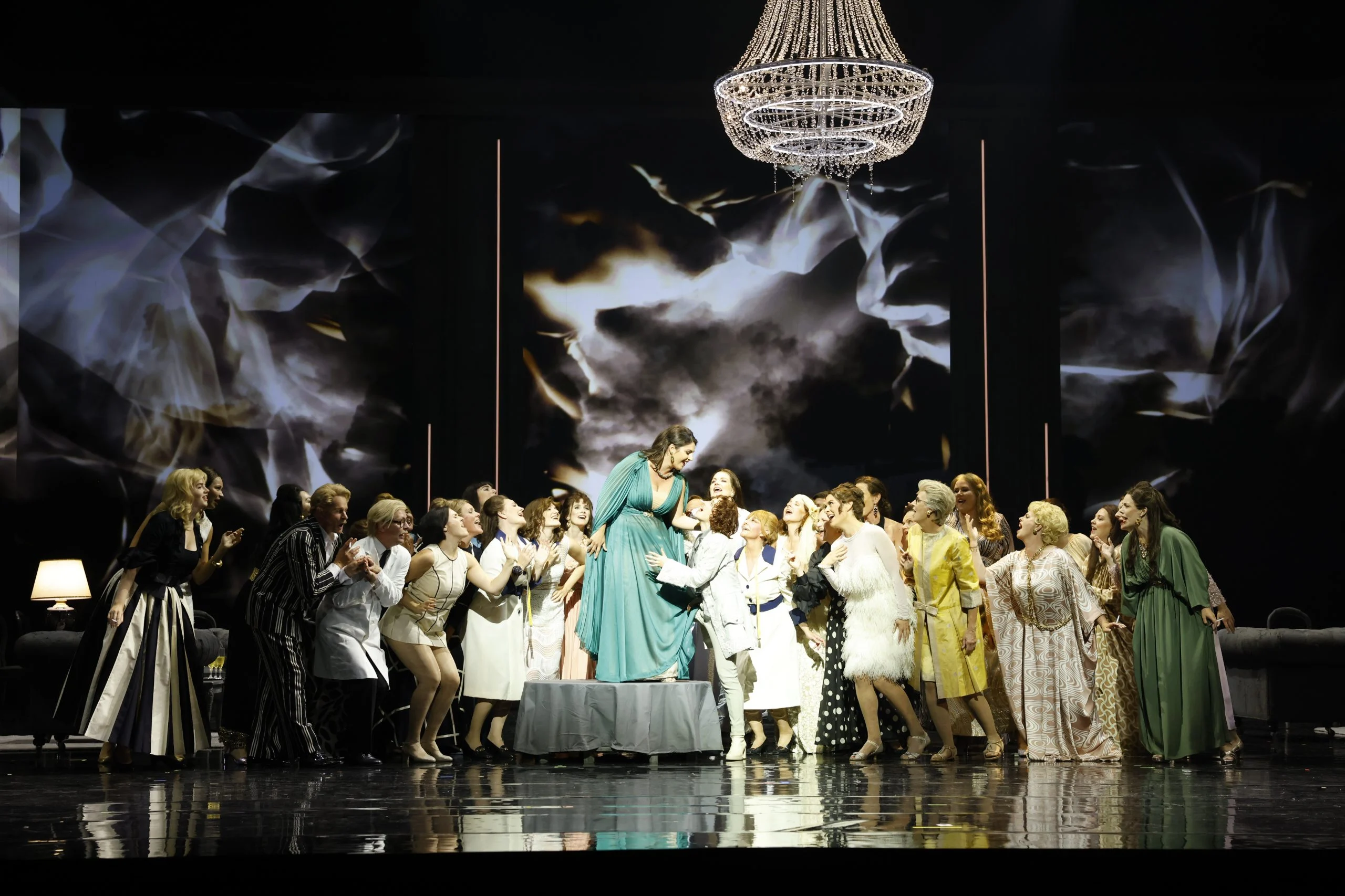 Coro femminile in abiti colorati intorno a una cantante in verde in piedi su un tavolo, sotto un lampadario di cristallo – Don Carlo al Teatro Reale Danese, Copenaghen, Danimarca, 2024 – scenografia d'opera