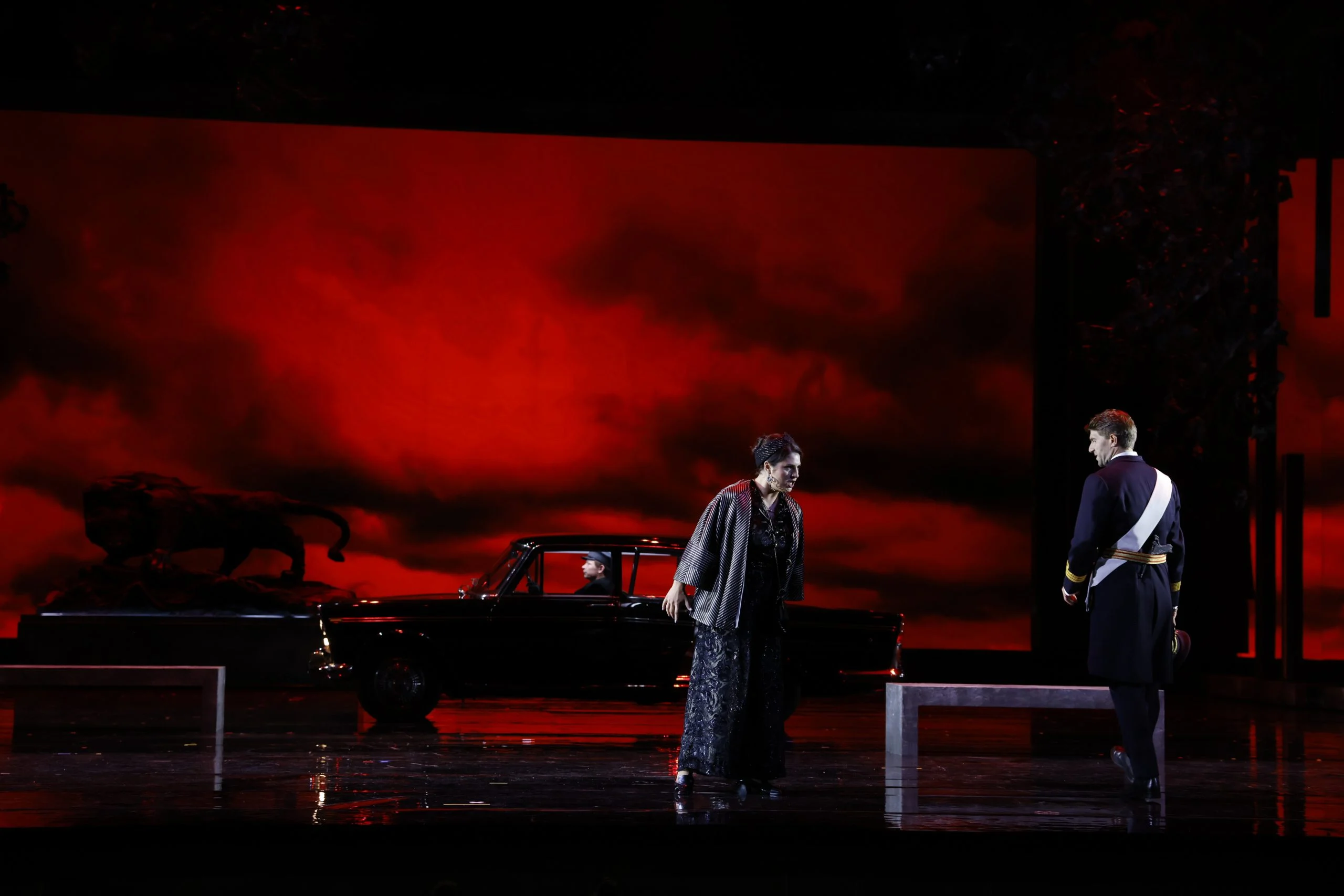 Cantante in nero davanti a una proiezione di cielo rosso fuoco, un ufficiale in uniforme e un'auto nera dietro – Don Carlo al Teatro Reale Danese, Copenaghen, Danimarca, 2024 – scenografia d'opera