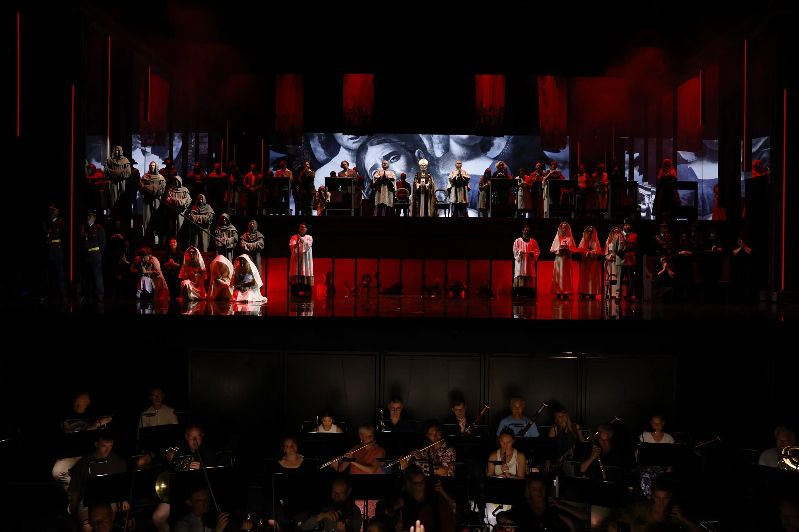 Penitenti incappucciati e coro su gradinate illuminate di rosso sopra la buca dell'orchestra, volti proiettati dietro – Don Carlo al Teatro Reale Danese, Copenaghen, Danimarca, 2024 – scenografia d'opera