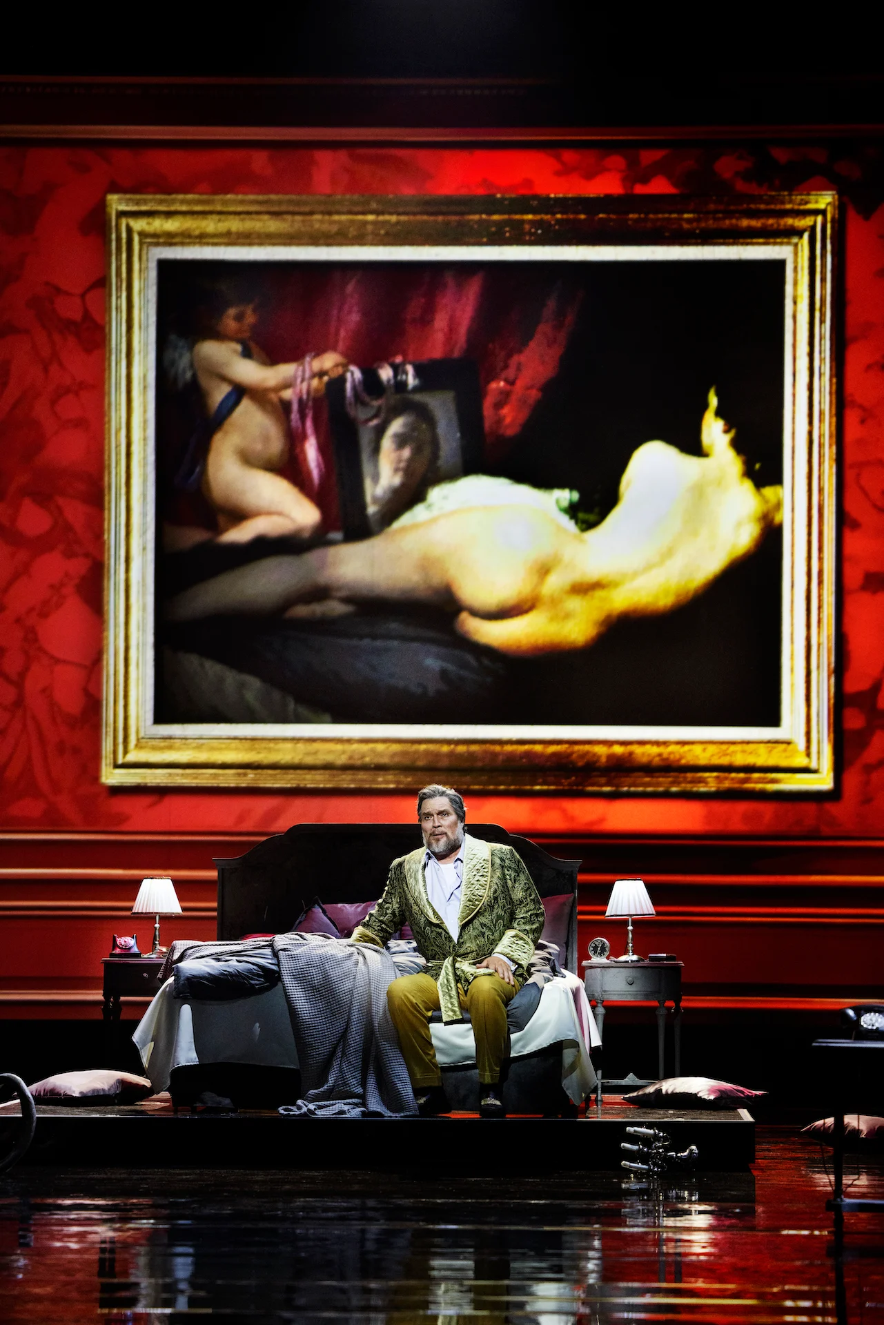 Cantante in una veste di broccato verde seduto su un letto sotto un enorme dipinto incorniciato di un nudo disteso – Don Carlo al Teatro Reale Danese, Copenaghen, Danimarca, 2024 – scenografia d'opera