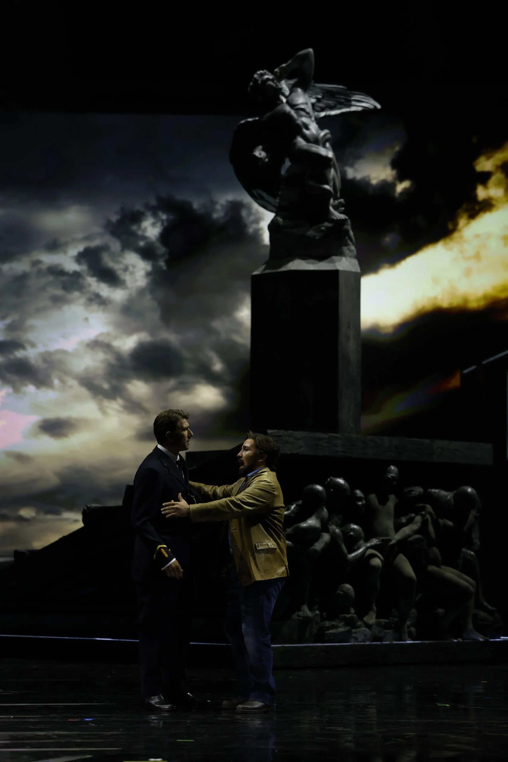 Due uomini uno di fronte all'altro sotto il monumento con la statua alata, contro la proiezione di un cielo in tempesta – Don Carlo al Teatro Reale Danese, Copenaghen, Danimarca, 2024 – scenografia d'opera