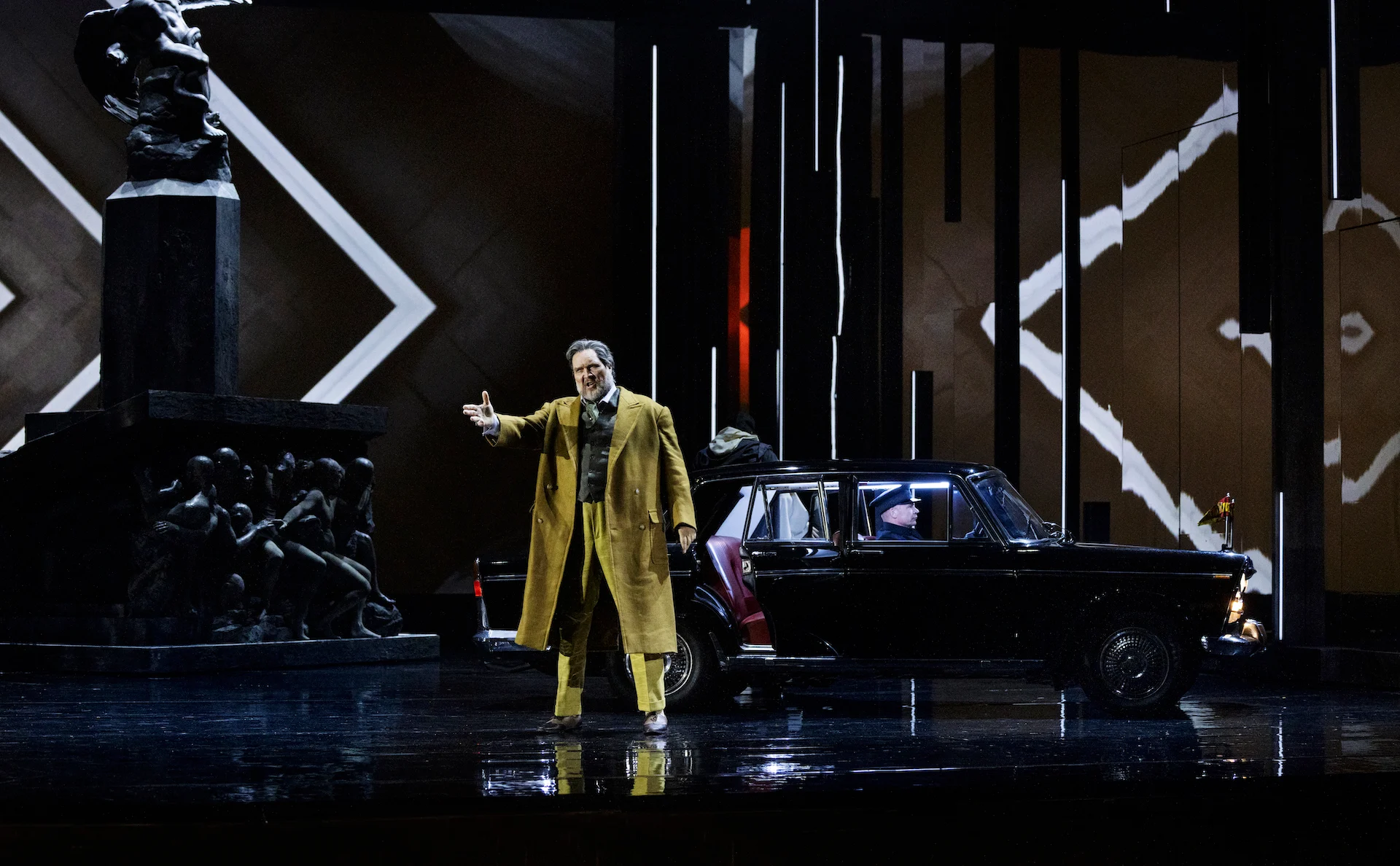 Cantante in soprabito giallo che gesticola accanto a un'auto d'epoca nera, il monumento e linee bianche a zigzag dietro – Don Carlo al Teatro Reale Danese, Copenaghen, Danimarca, 2024 – scenografia d'opera
