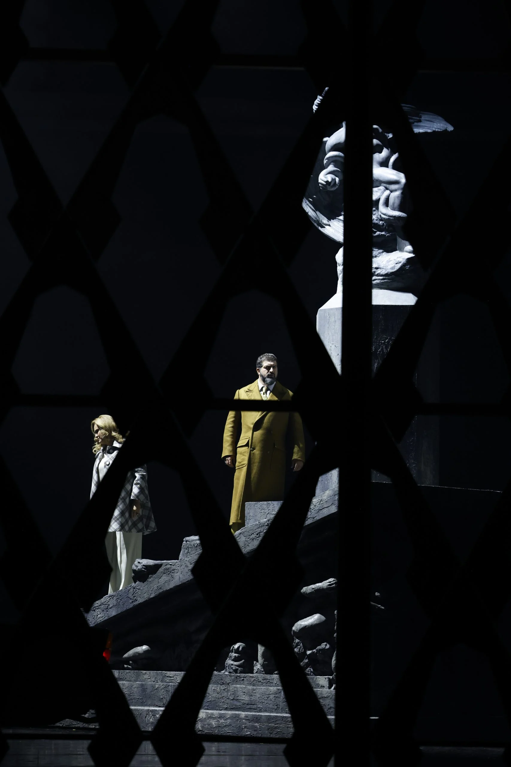 Due cantanti sui gradini del monumento visti attraverso un reticolo scuro a losanghe in primo piano – Don Carlo al Teatro Reale Danese, Copenaghen, Danimarca, 2024 – scenografia d'opera