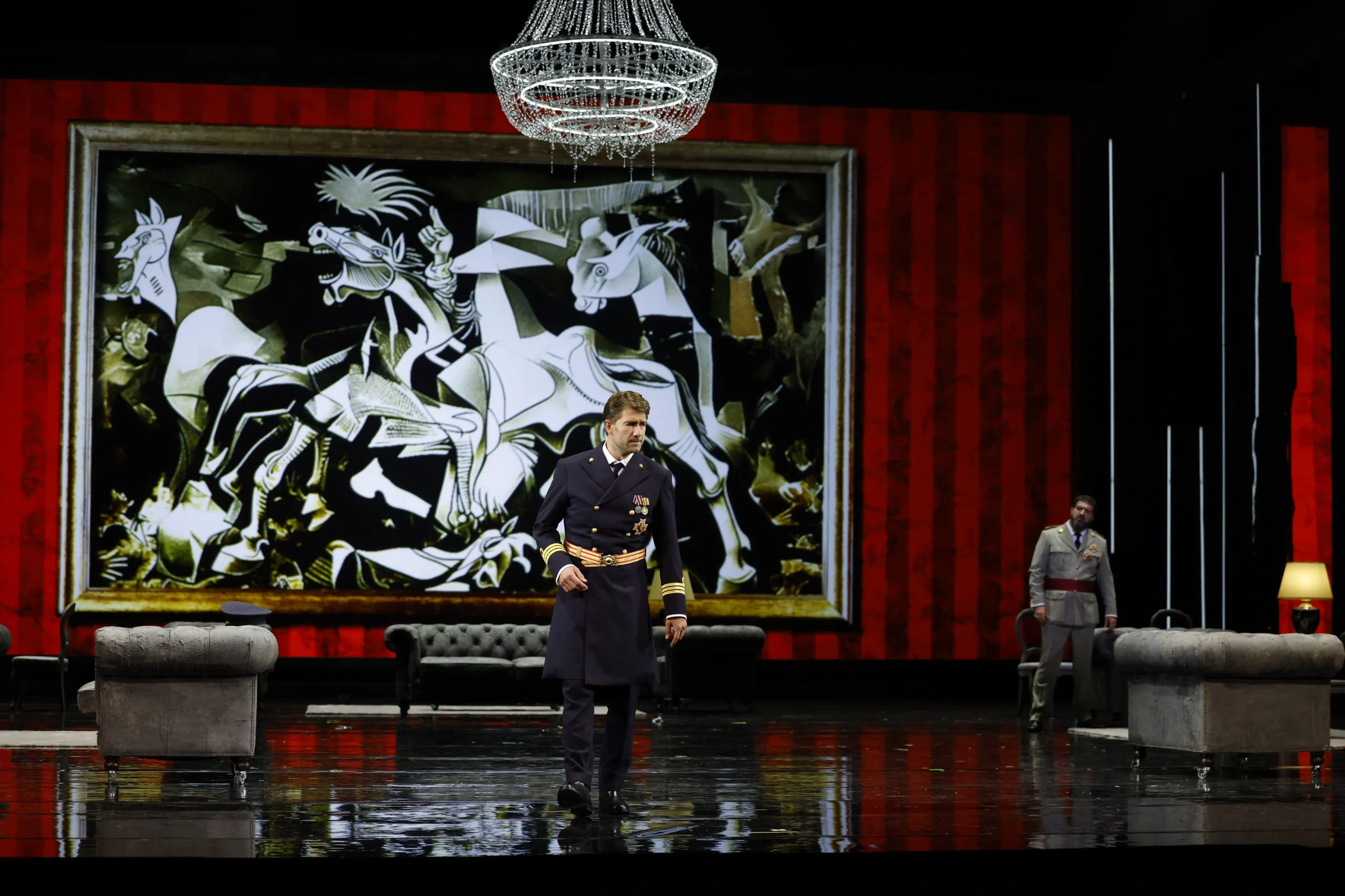 Ufficiale in uniforme della marina davanti a un'enorme riproduzione di Guernica di Picasso, un lampadario e tende rosse – Don Carlo al Teatro Reale Danese, Copenaghen, Danimarca, 2024 – scenografia d'opera
