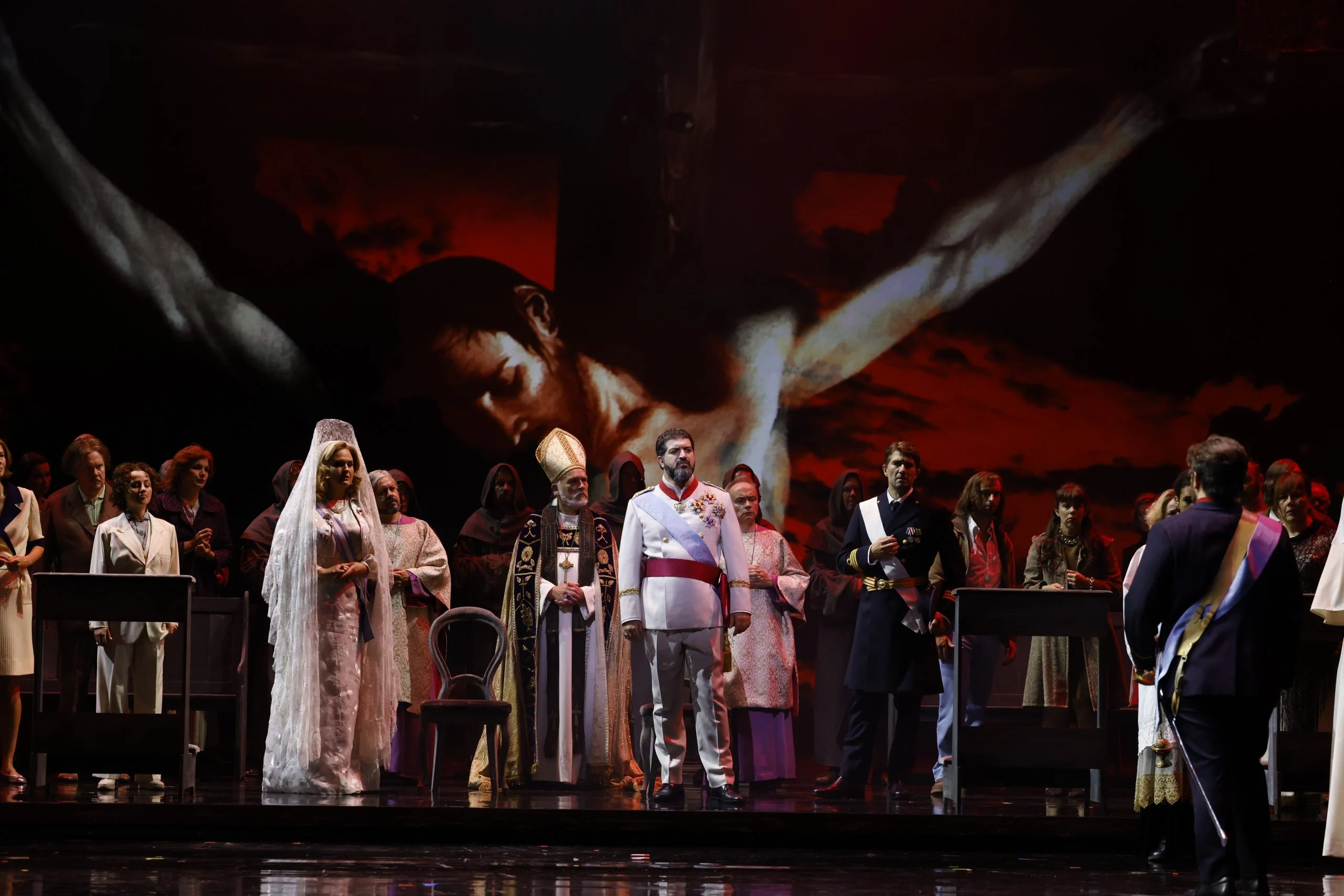 Sposa con velo bianco, un vescovo, un ufficiale in uniforme bianca e il coro davanti a un gigantesco crocifisso proiettato – Don Carlo al Teatro Reale Danese, Copenaghen, Danimarca, 2024 – scenografia d'opera