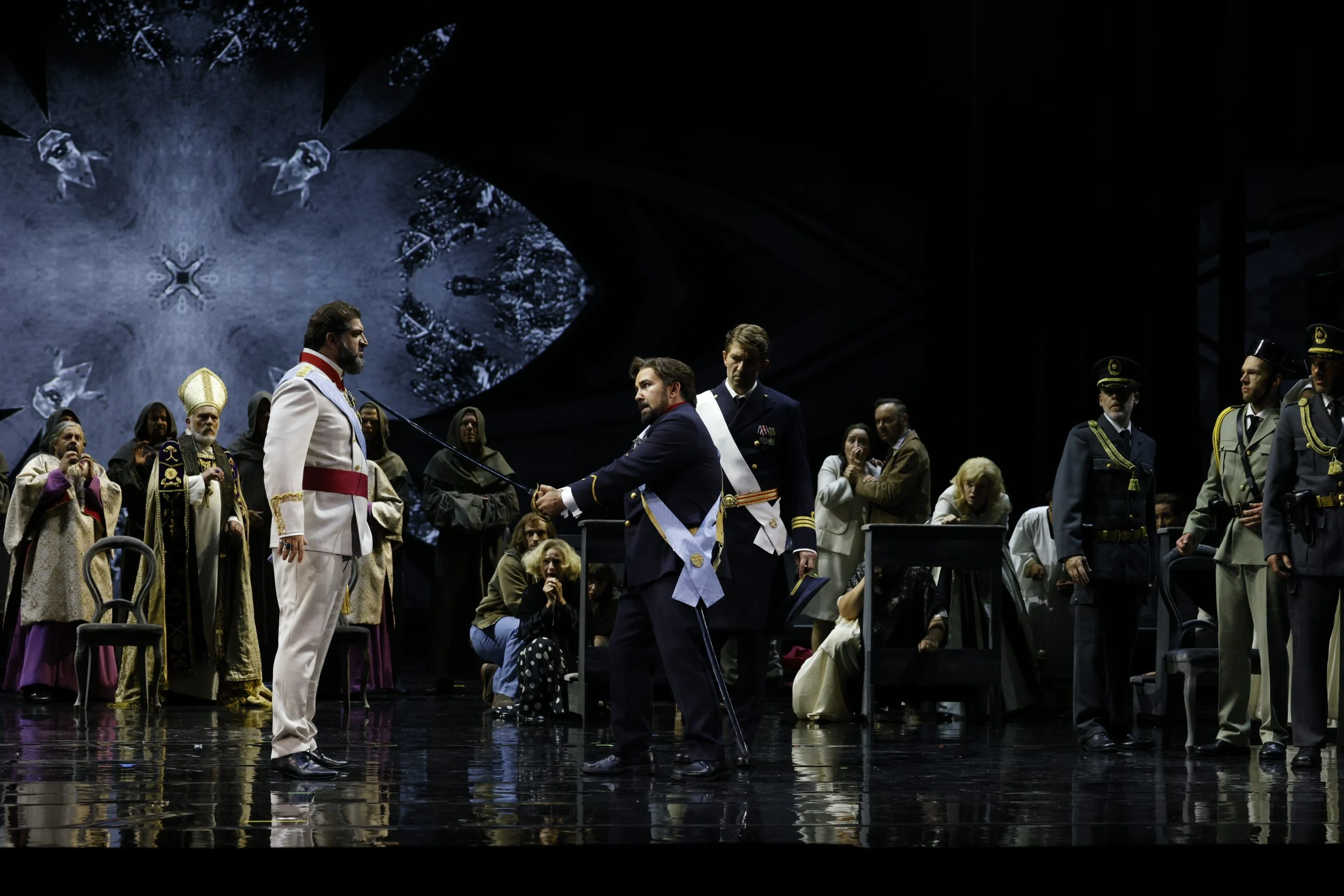 Scontro tra uomini in uniforme e fusciacca, con clero e coro che osservano, un rosone proiettato dietro – Don Carlo al Teatro Reale Danese, Copenaghen, Danimarca, 2024 – scenografia d'opera