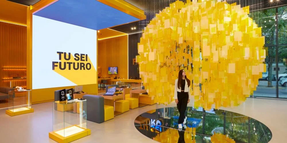Visitatore sotto la sfera sospesa di tessere gialle, riflesso in uno specchio rotondo nel pavimento – STEP FuturAbility District by Fastweb, Milano, Italia, 2022 – allestimento espositivo