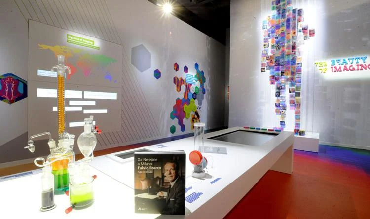 Sala espositiva con vetreria da laboratorio e un libro su un tavolo bianco, dietro una figura di immagini colorate – Bracco The Beauty of Imaging, Milano, Italia, 2017 – allestimento espositivo