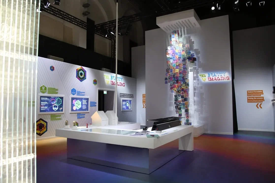 Vista d'insieme della sala espositiva con un tavolo bianco centrale, grafiche alle pareti e la figura di immagini sospesa – Bracco The Beauty of Imaging, Milano, Italia, 2017 – allestimento espositivo