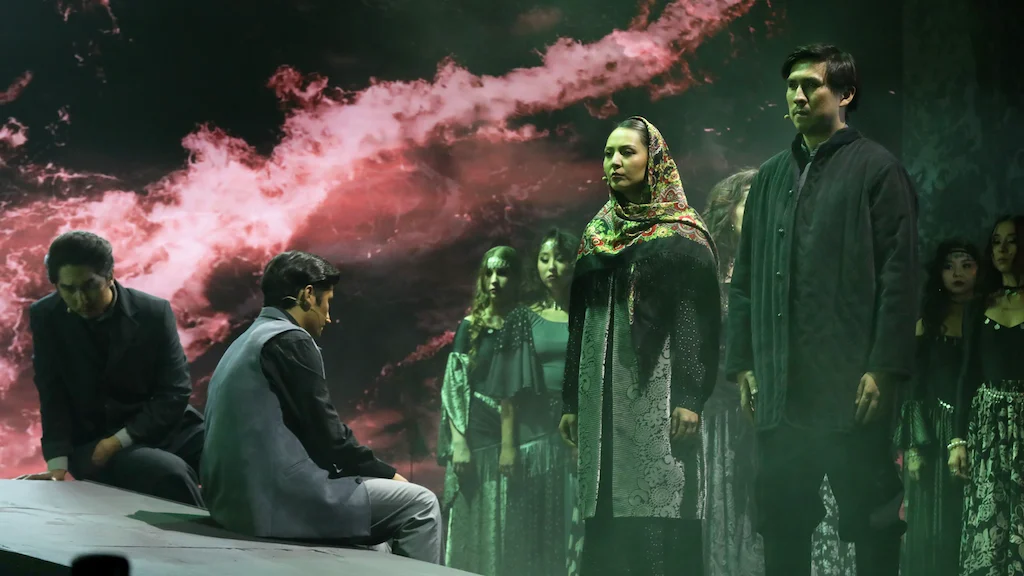 Artisti in abiti tradizionali davanti a una proiezione di fumo rosso, due uomini seduti sul bordo del palco – Waltz Karoly al Turkistan Theatre, Kazakistan, 2024 – scenografia