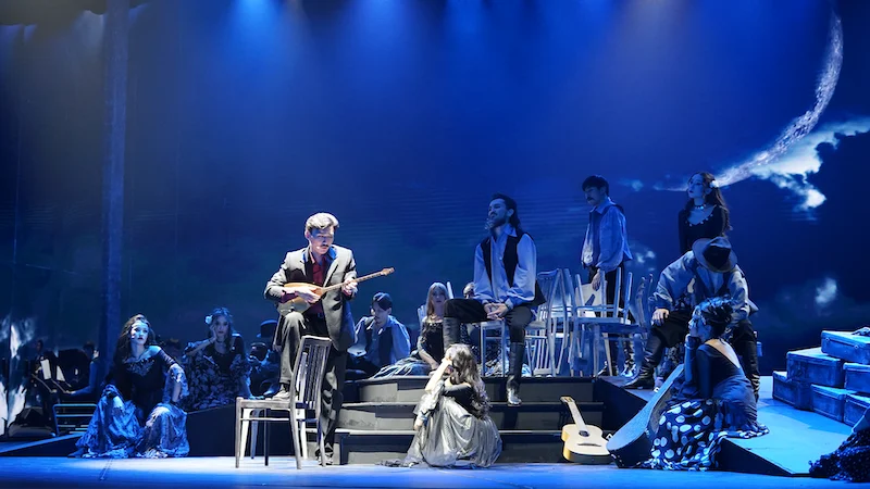 Scena notturna blu con la proiezione di una falce di luna, un suonatore di mandolino seduto tra gli artisti sui gradini – Waltz Karoly al Turkistan Theatre, Kazakistan, 2024 – scenografia