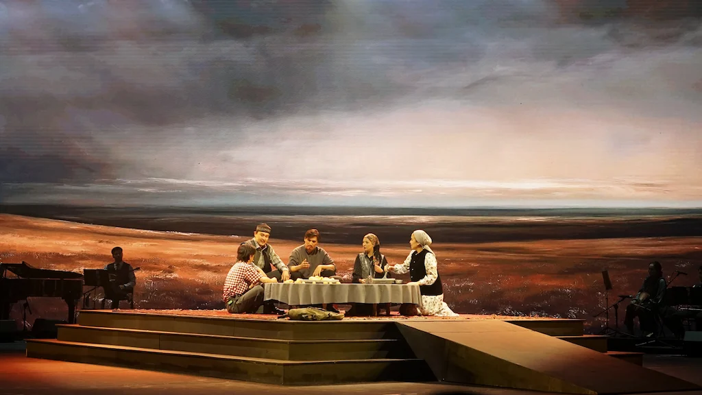 Scena d'opera: famiglia seduta a tavola su una pedana a gradoni davanti alla proiezione di una vasta steppa – Waltz Karoly al Turkistan Theatre, Kazakistan, 2024 – scenografia