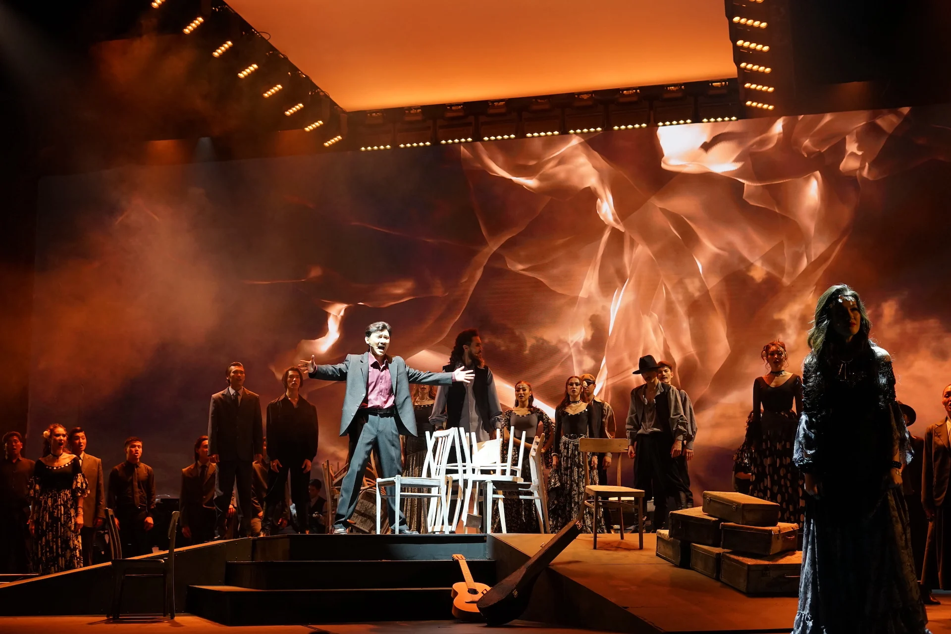 Scena d'insieme con sedie bianche e casse impilate, un cantante a braccia aperte davanti a proiezioni arancioni vorticose – Waltz Karoly al Turkistan Theatre, Kazakistan, 2024 – scenografia