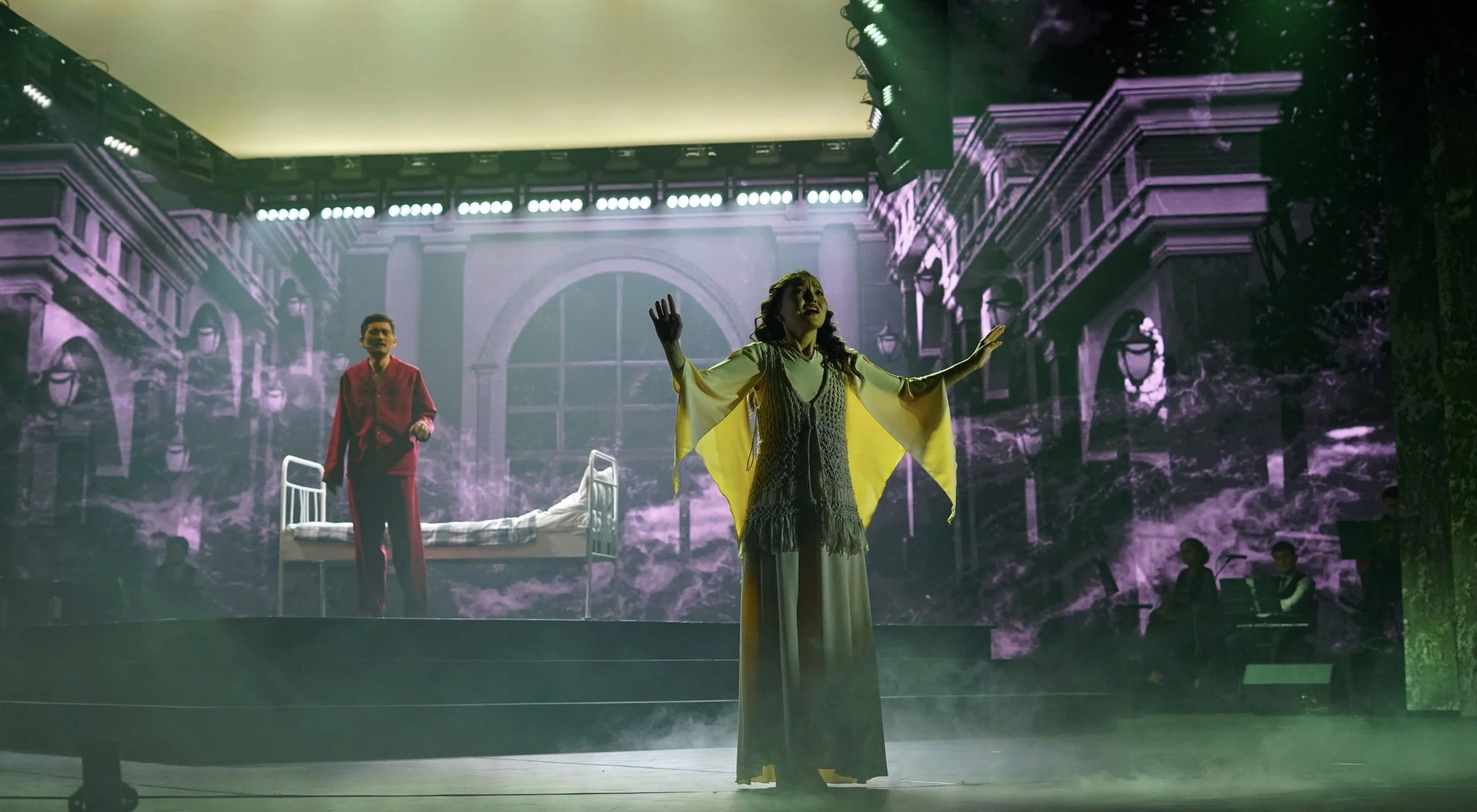 Cantante in mantello giallo al centro del palco, un letto d'ospedale dietro e facciate di palazzi proiettate tutto intorno – Waltz Karoly al Turkistan Theatre, Kazakistan, 2024 – scenografia
