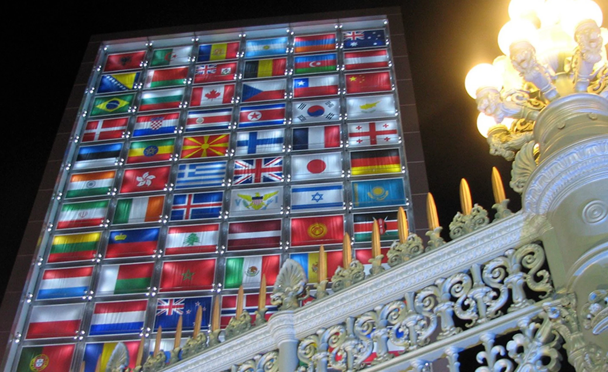 Parete illuminata di bandiere nazionali di notte, vista dietro una balaustra bianca decorata e un lampione – Medals Plaza, Olimpiadi invernali di Torino 2006, Italia – progettazione di eventi