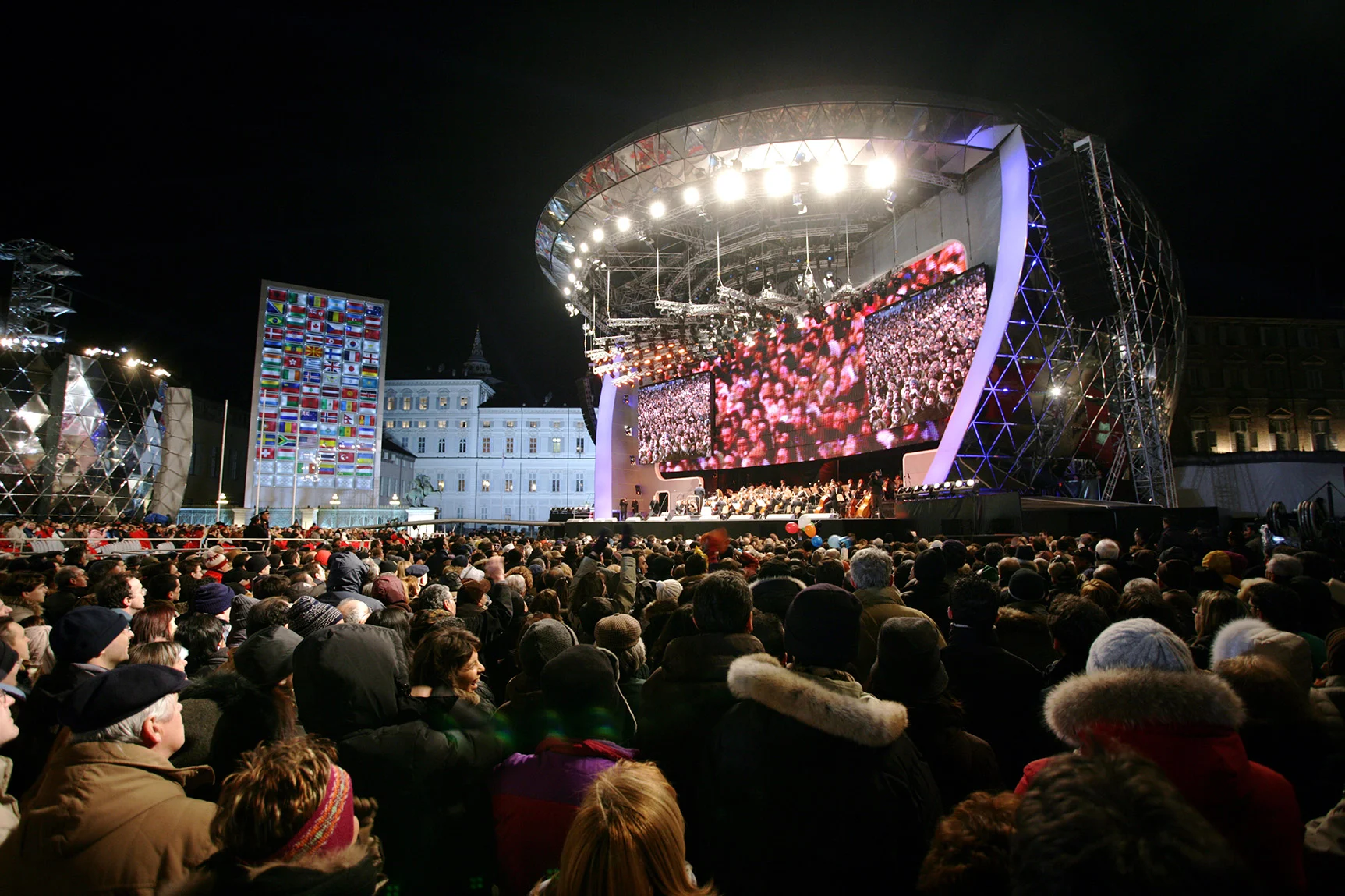 Medals Plaza in Piazza Castello di notte: palco curvo con schermi giganti, una parete di bandiere e una grande folla – Medals Plaza, Olimpiadi invernali di Torino 2006, Italia – progettazione di eventi
