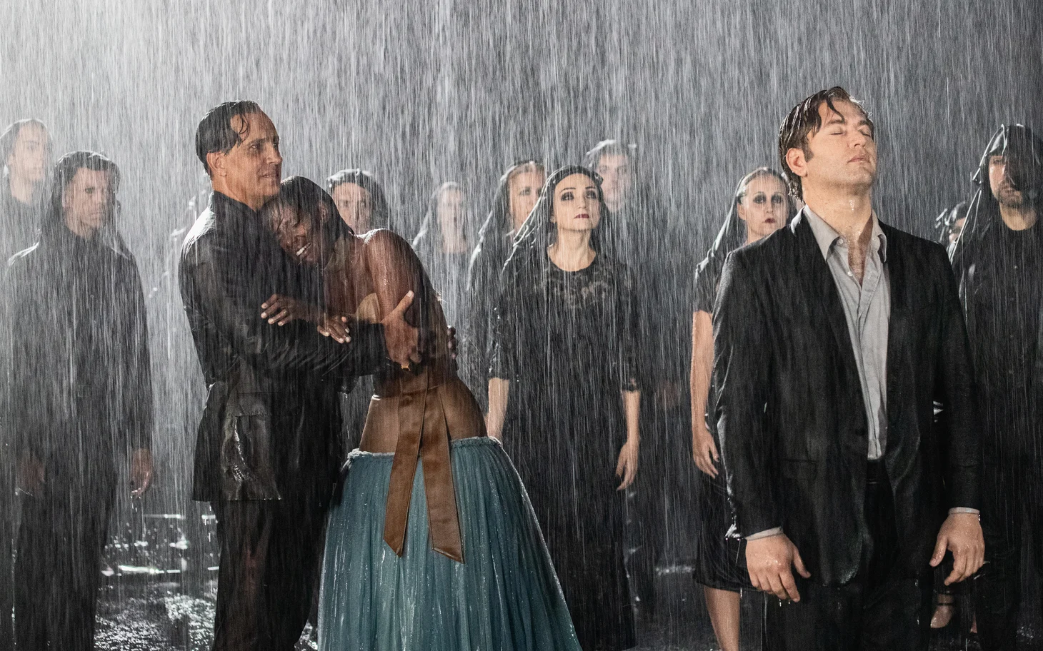 Fotogramma del film: una coppia abbracciata e un uomo in abito scuro tra la folla sotto una pioggia battente – The Opera! Arie per un'Eclissi alla Festa del Cinema di Roma, Italia, 2024 – scenografia