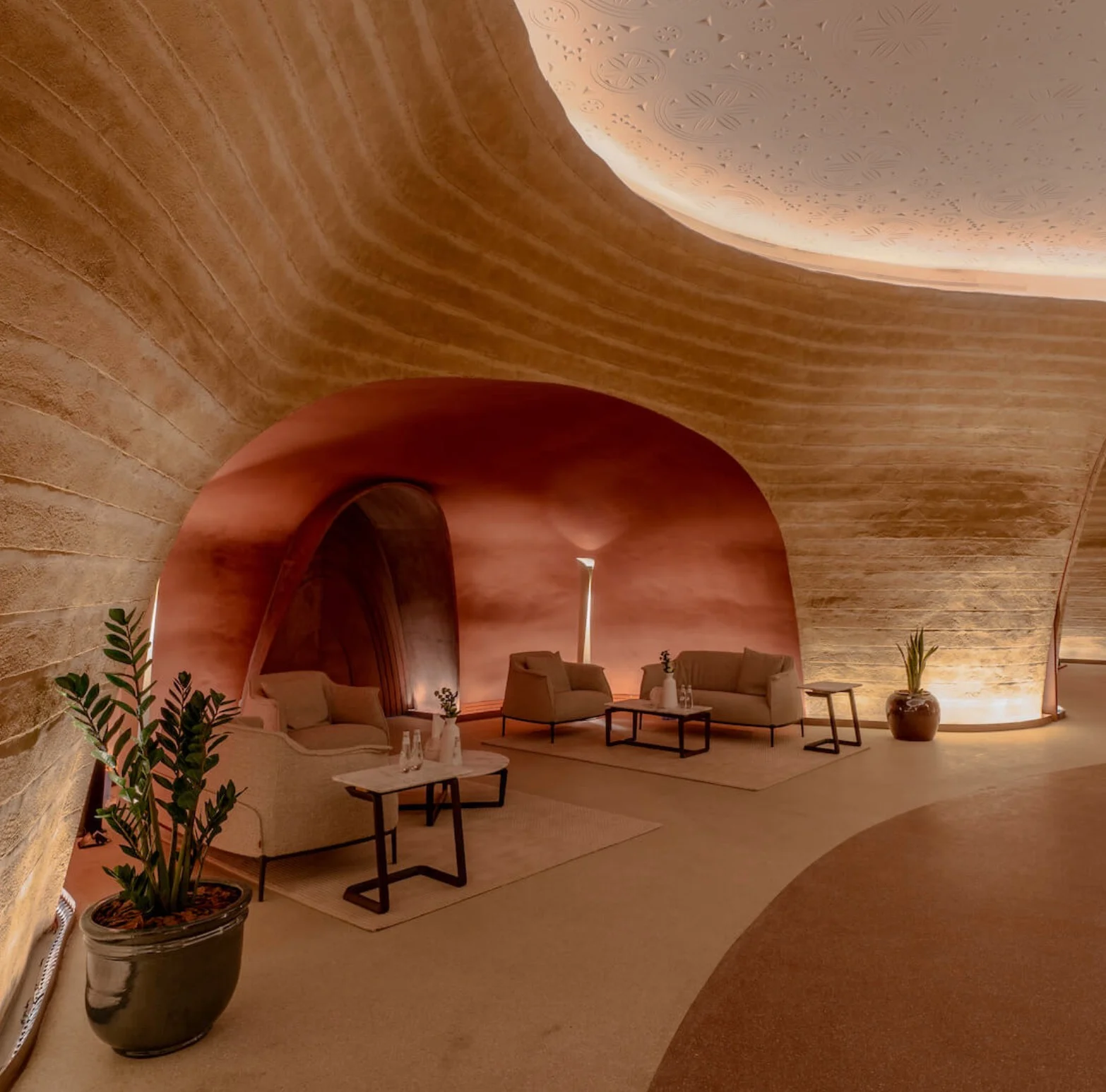 Lounge in terra cruda con poltrone e tavolini bassi sotto un arco in terracotta nel centro culturale – King Fahad Chair per Poltrona Frau, Riyadh, Arabia Saudita, 2024 – design di prodotto