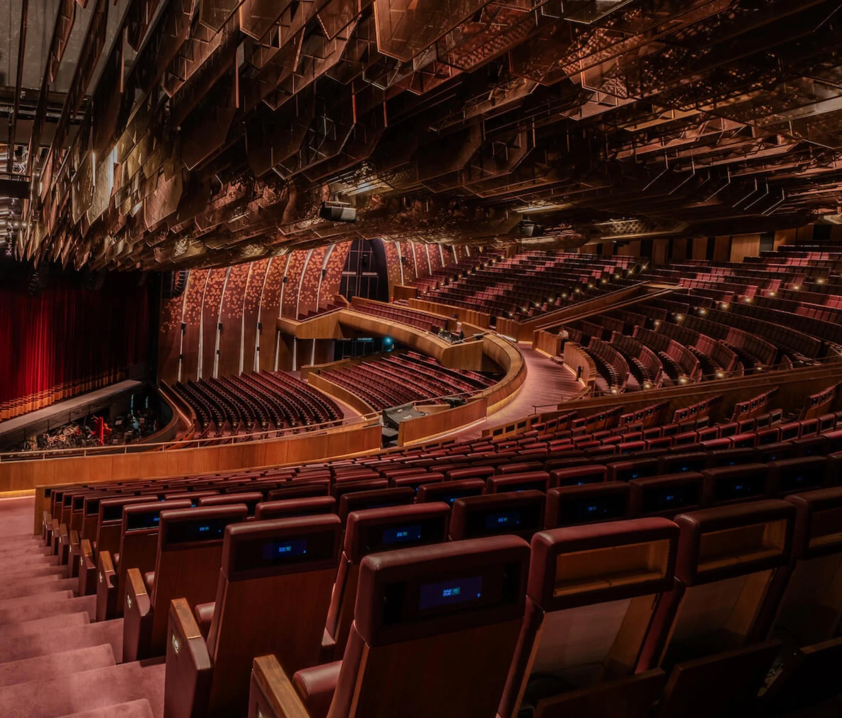 Interno del teatro dal corridoio laterale: poltrone rosse a gradinata e balconate sotto un soffitto sfaccettato in rame – King Fahad Chair per Poltrona Frau, Riyadh, Arabia Saudita, 2024 – design di prodotto