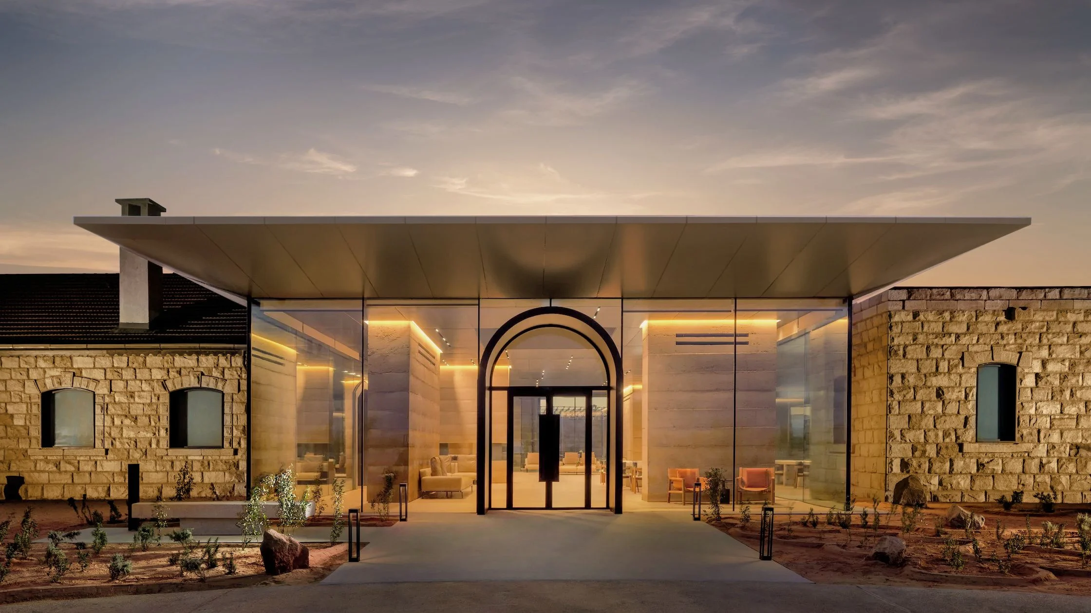 Ingresso dell'hotel al tramonto: un padiglione in vetro sotto un sottile tetto piano tra due antichi edifici in pietra – The Chedi Hegra, AlUla, Arabia Saudita, 2024 – architettura alberghiera