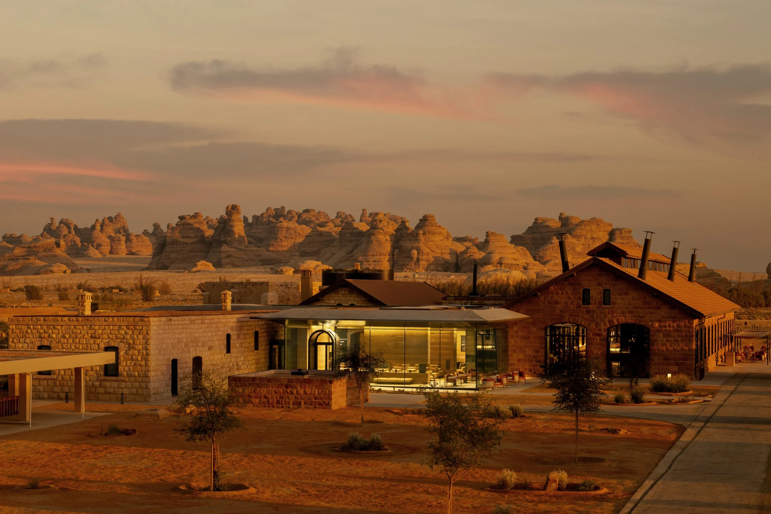 The Chedi Hegra al tramonto: edifici in pietra restaurati e un padiglione in vetro davanti alle formazioni rocciose di AlUla – The Chedi Hegra, AlUla, Arabia Saudita, 2024 – architettura alberghiera