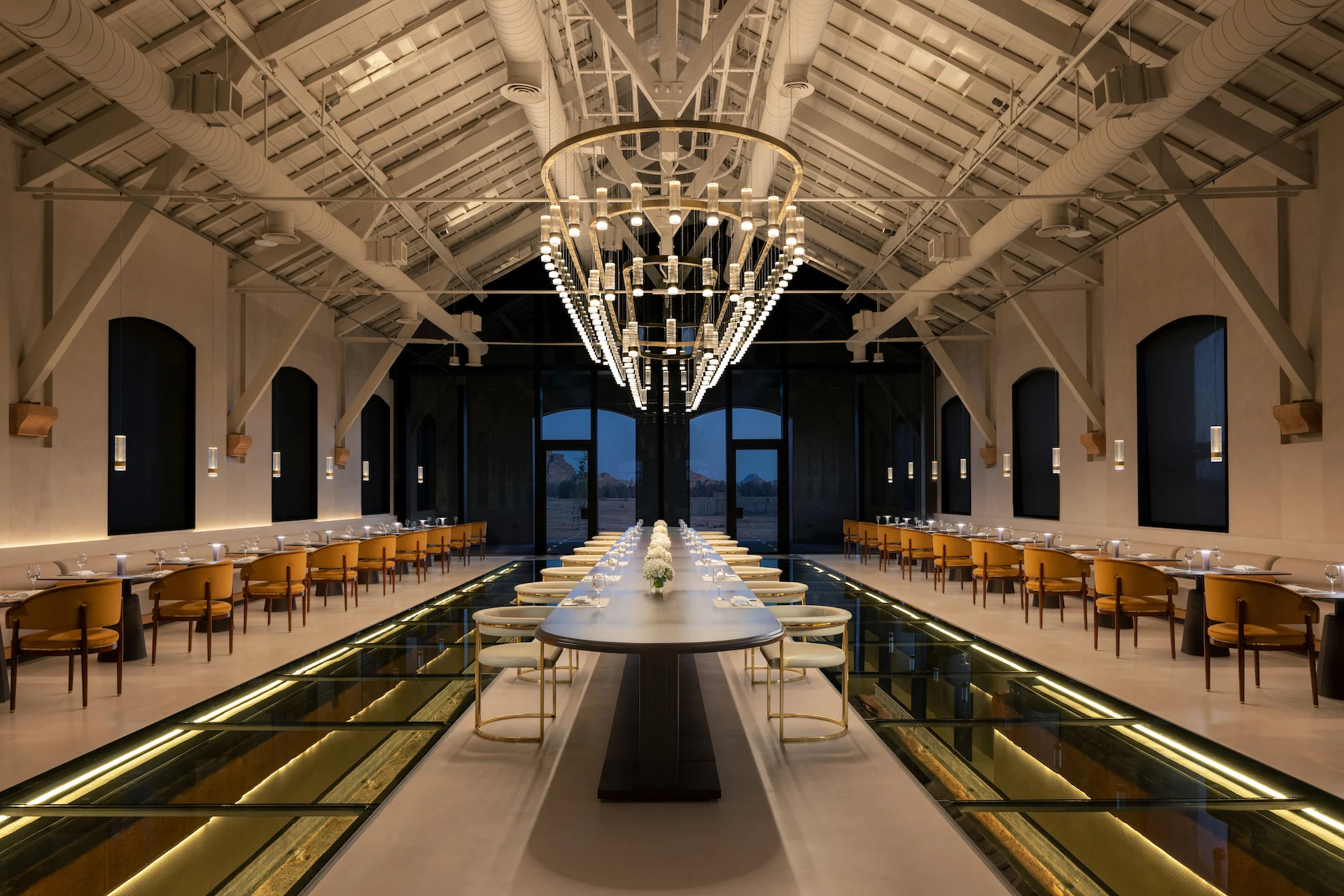 Ristorante in una sala restaurata: lungo tavolo sotto un lampadario ad anello, pannelli di pavimento in vetro e capriate a vista – The Chedi Hegra, AlUla, Arabia Saudita, 2024 – architettura alberghiera