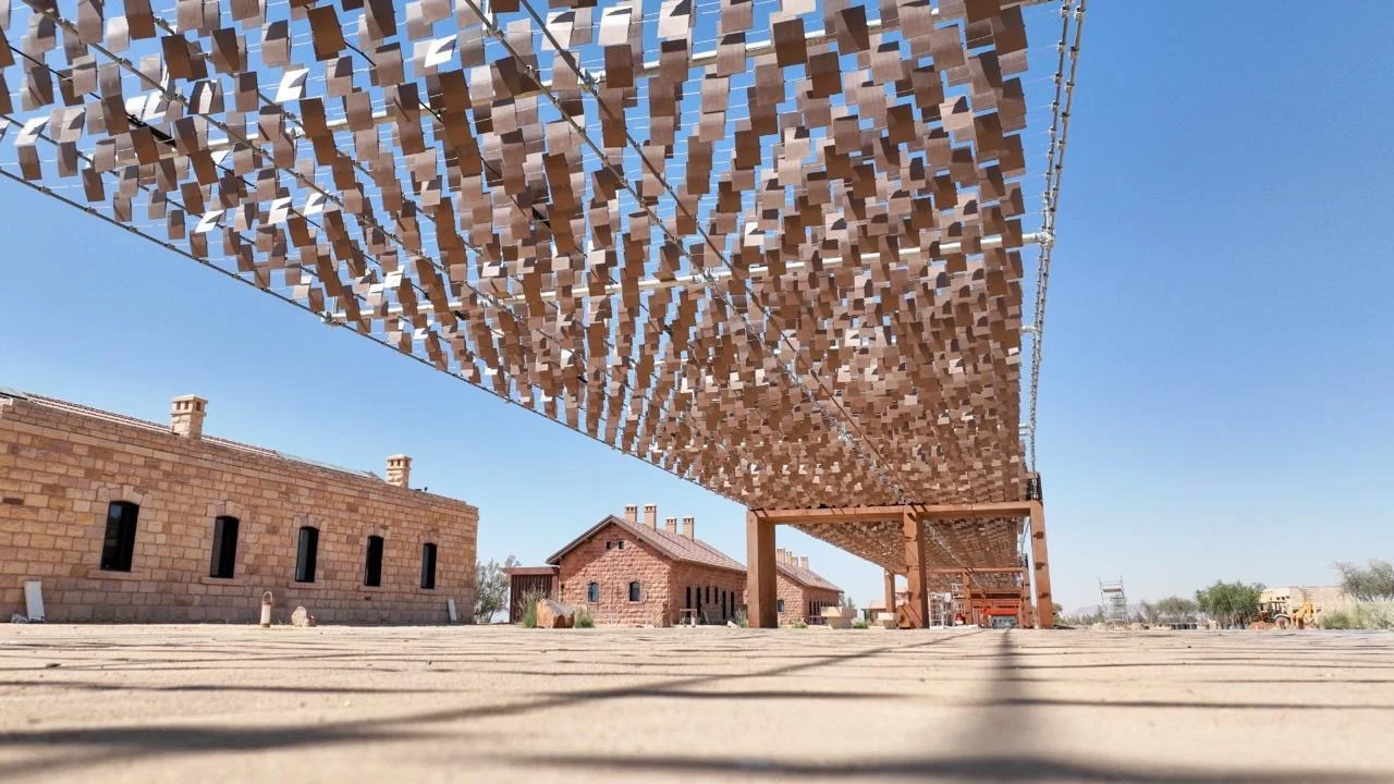 Vista dal basso delle migliaia di piccoli elementi metallici della pensilina contro il cielo blu, accanto a case in pietra – The Chedi Hegra Art Canopy, AlUla, Arabia Saudita, 2025 – architettura