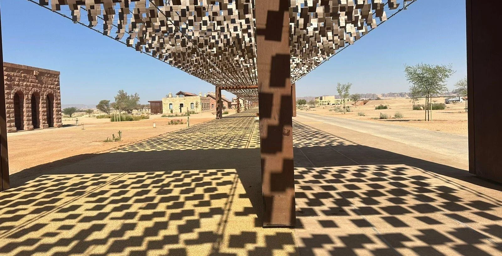 Ombre a motivi proiettate a terra dalla copertura cinetica, con edifici storici in pietra e il deserto sullo sfondo – The Chedi Hegra Art Canopy, AlUla, Arabia Saudita, 2025 – architettura