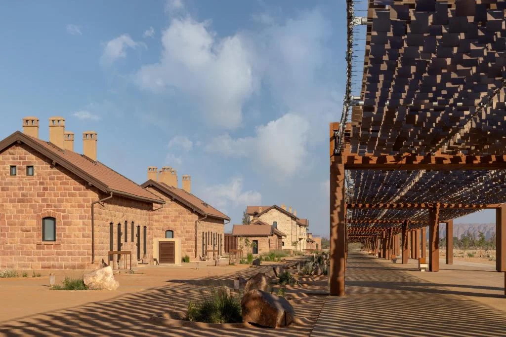 Copertura su pali color ruggine lungo edifici storici in arenaria, con le sue ombre che disegnano strisce sulla sabbia – The Chedi Hegra Art Canopy, AlUla, Arabia Saudita, 2025 – architettura