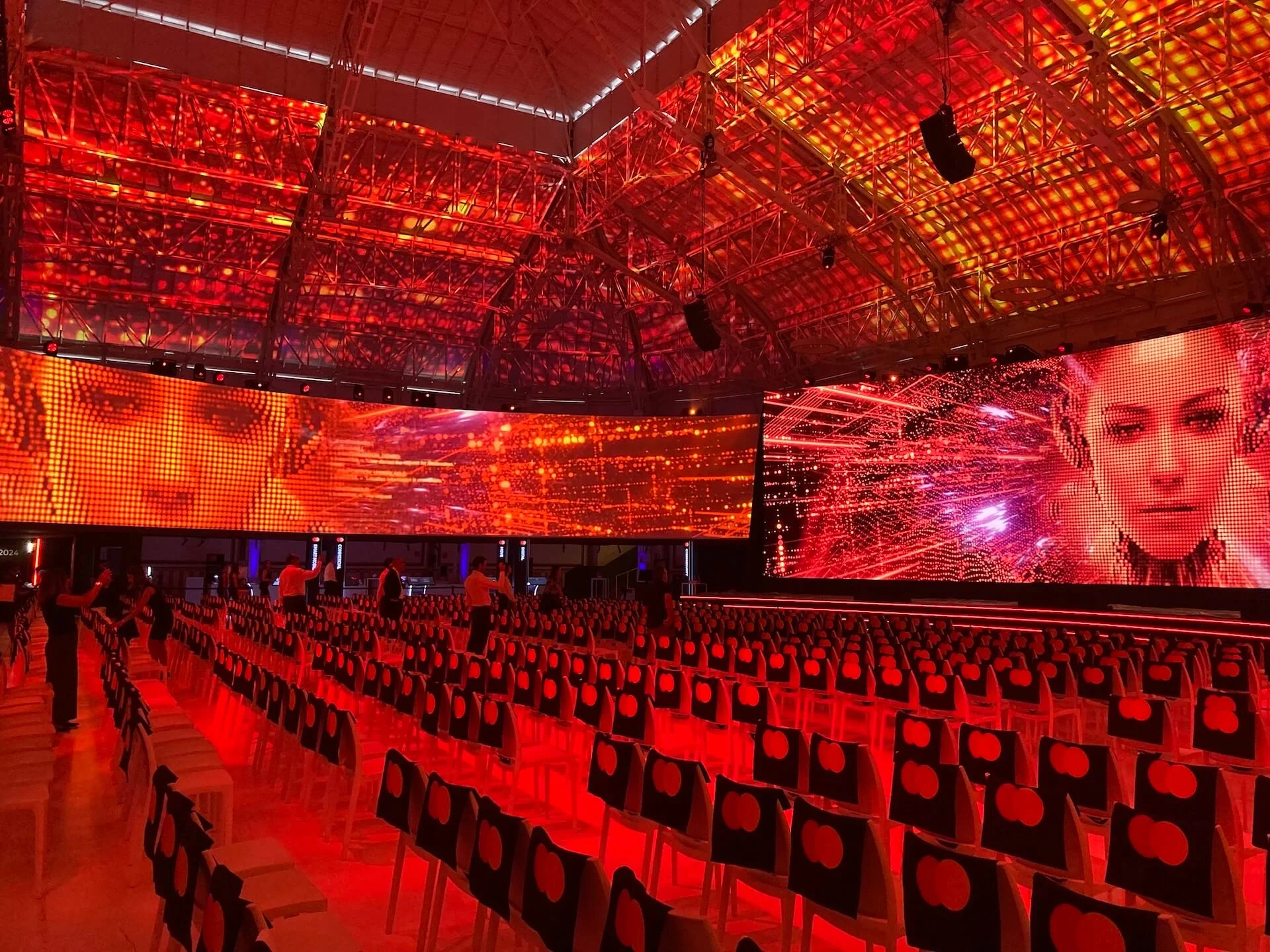 Sala illuminata di rosso con schermi LED che mostrano volti femminili sopra file di sedie con il logo Mastercard – Mastercard Innovation Forum 2024, Milano, Italia – progettazione di eventi