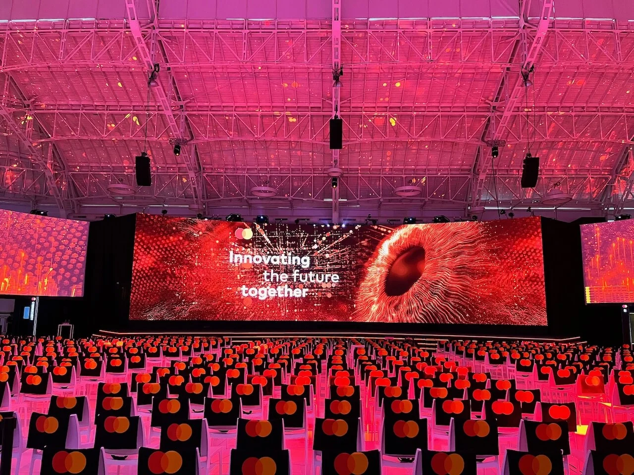 Schermo del palco con la scritta Innovating the future together su una grafica di un occhio rosso, in una sala illuminata di rosa – Mastercard Innovation Forum 2024, Milano, Italia – progettazione di eventi