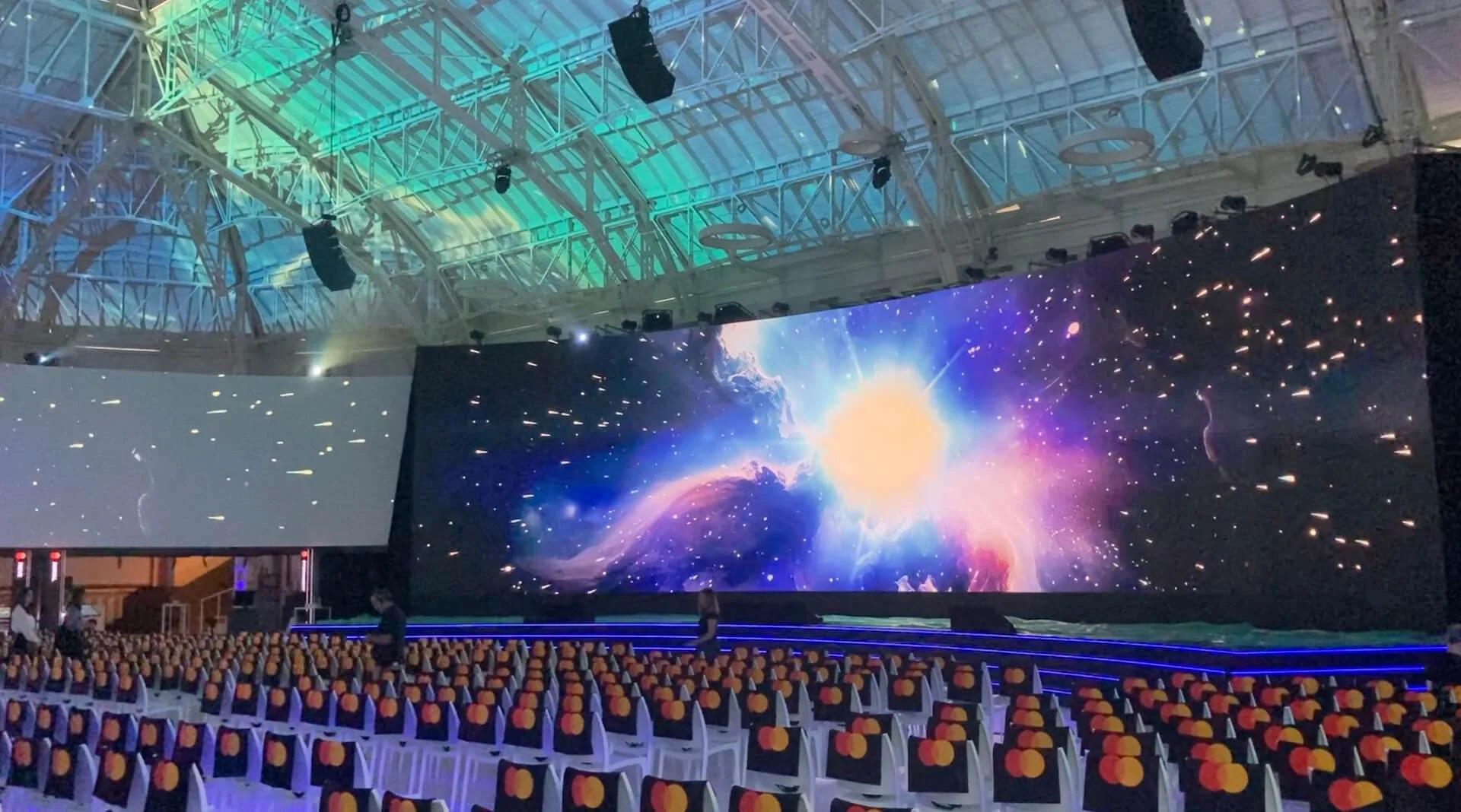 Schermo LED principale con un'esplosione cosmica luminosa sopra file di sedie, sotto la copertura in acciaio della sala – Mastercard Innovation Forum 2024, Milano, Italia – progettazione di eventi