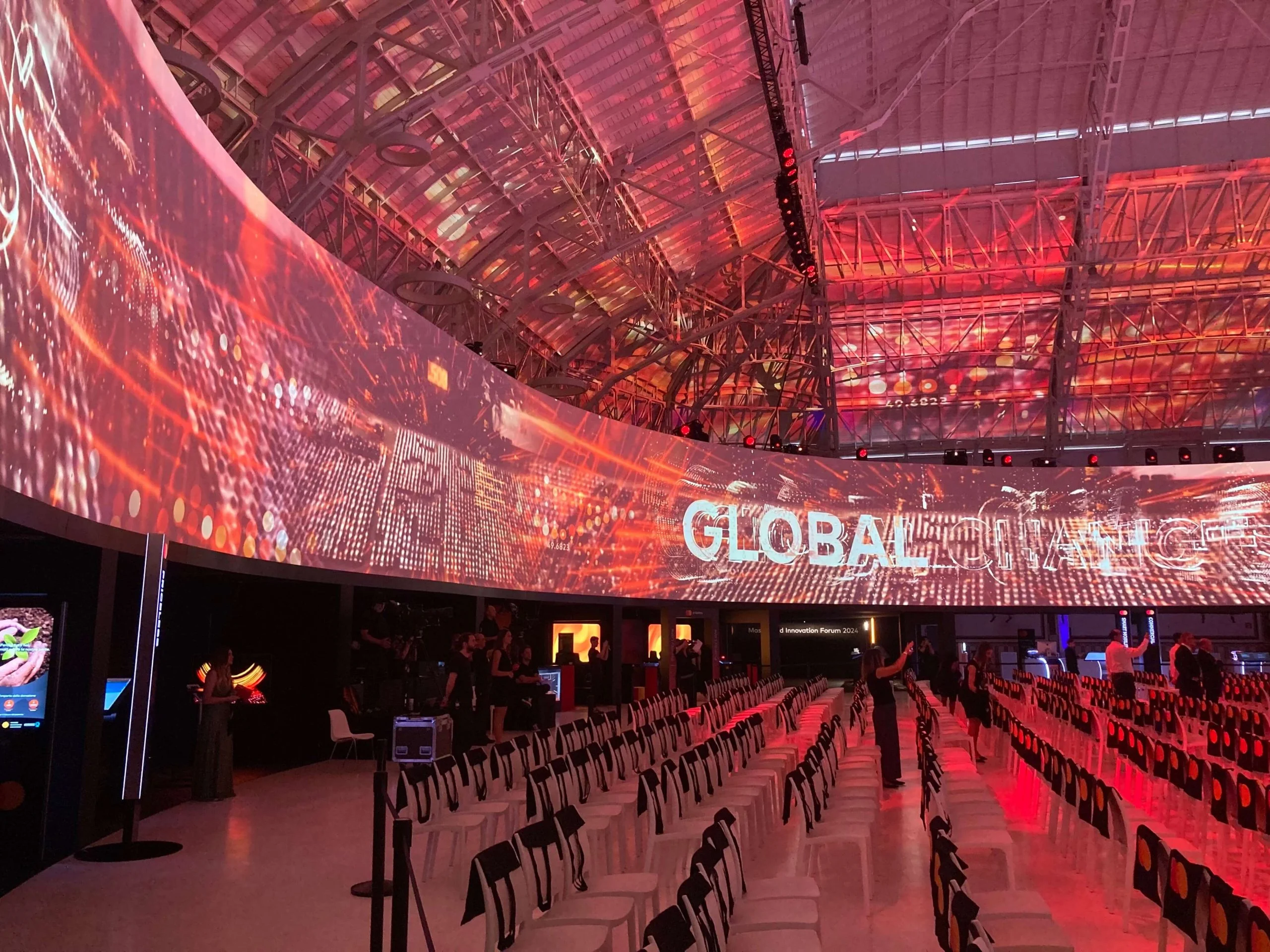 Parete LED curva che avvolge la sala con grafiche digitali rosse e la parola Global, sedie sotto – Mastercard Innovation Forum 2024, Milano, Italia – progettazione di eventi
